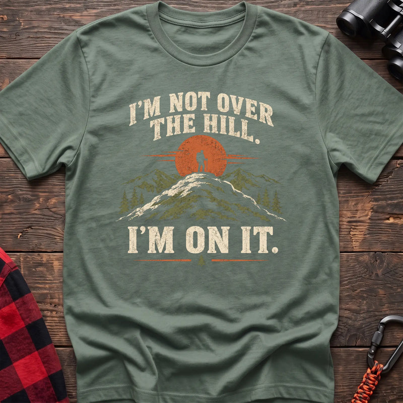 Sage cotton t-shirt with a hiker on a summit graphic and I’m Not Over the Hill I’m On It text on rustic wood