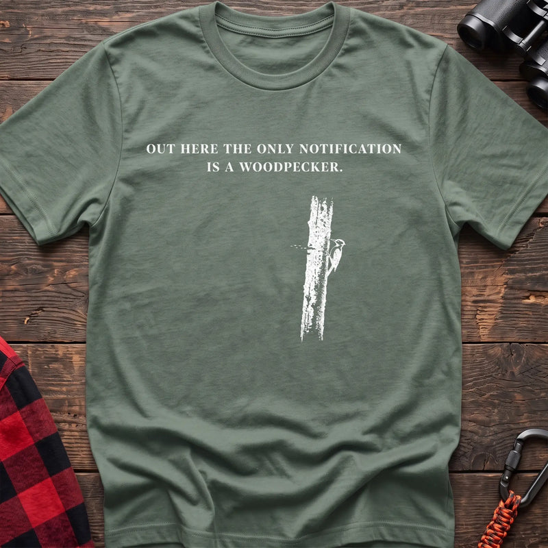 Sage cotton t-shirt with a woodpecker graphic and Out Here the Only Notification Is a Woodpecker text on rustic wood