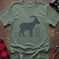 Sage green t-shirt with a dark silhouette of a mountain goat filled with topographic contour lines, and the vertical text “HIGHER GROUND” printed beside it, laid flat on a wooden surface.