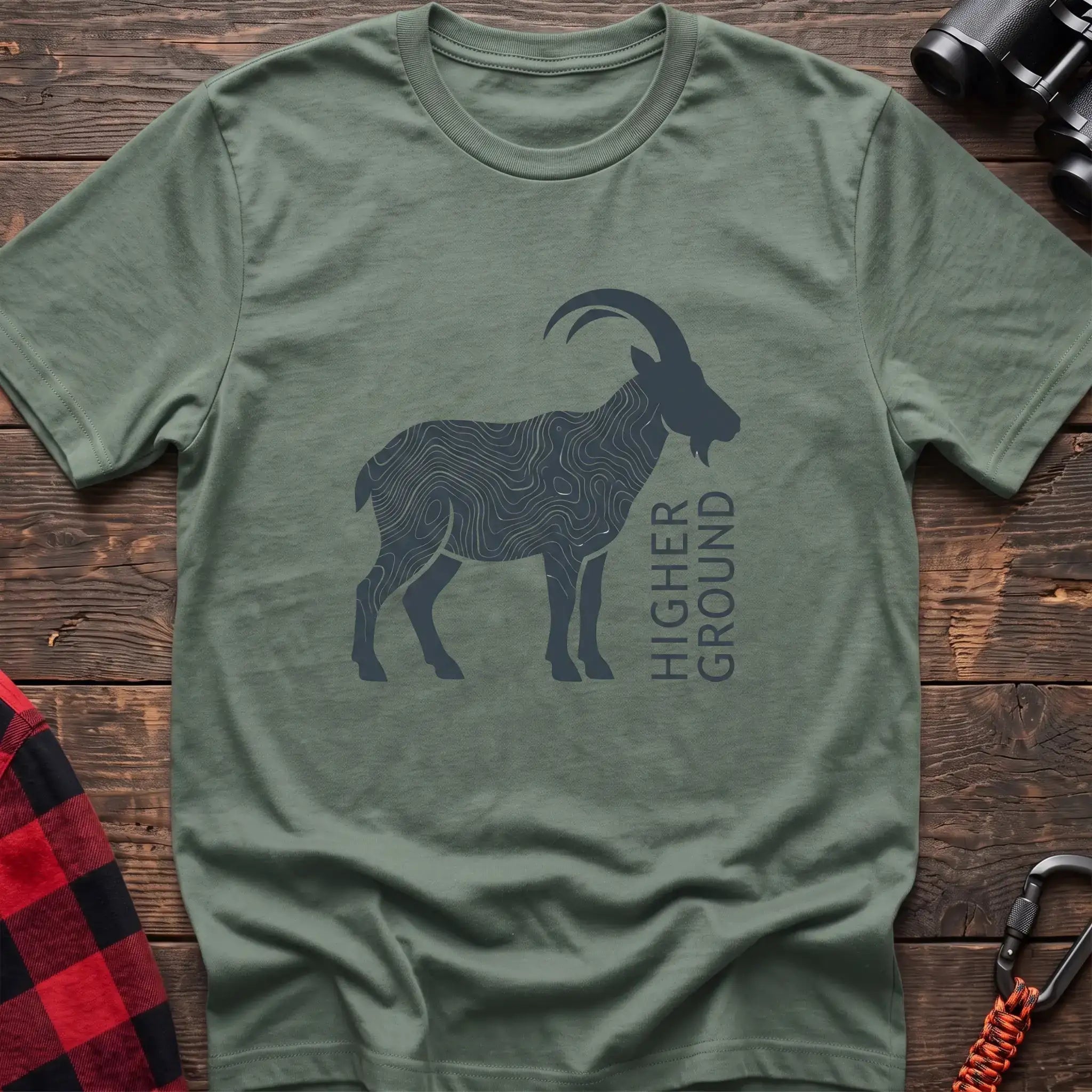 Sage green t-shirt with a dark silhouette of a mountain goat filled with topographic contour lines, and the vertical text “HIGHER GROUND” printed beside it, laid flat on a wooden surface.