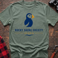 Sage cotton t-shirt with a navy puffin logo, yellow beak, and Rocky Shore Society Acadia text on a rustic wood background