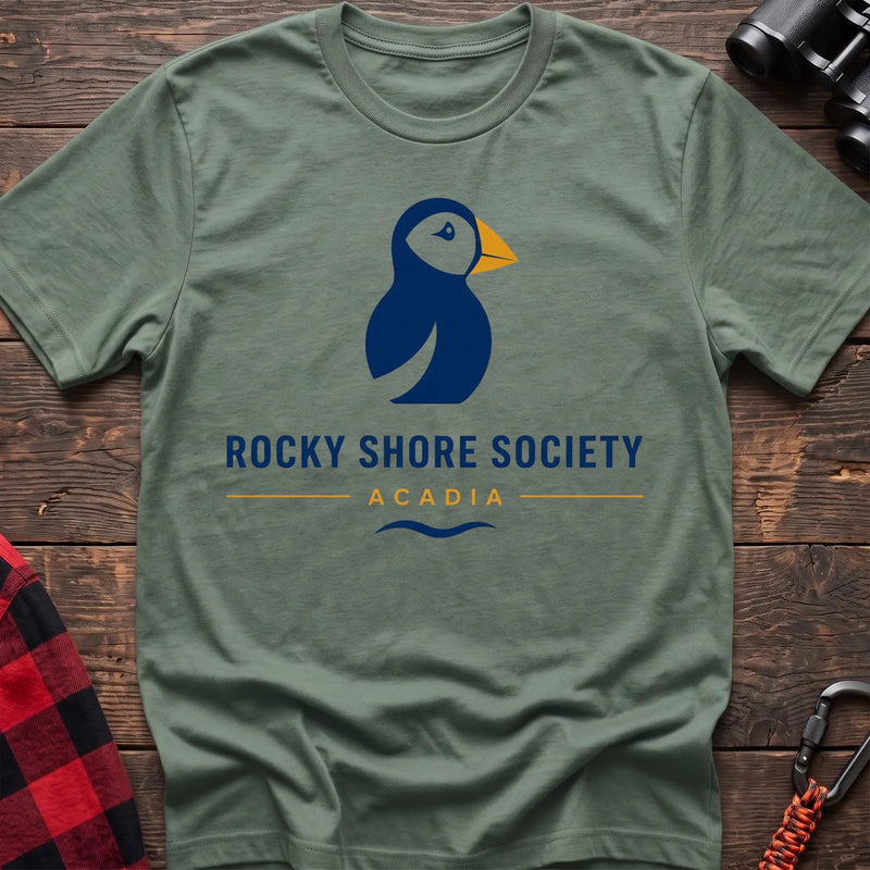 Sage cotton t-shirt with a navy puffin logo, yellow beak, and Rocky Shore Society Acadia text on a rustic wood background
