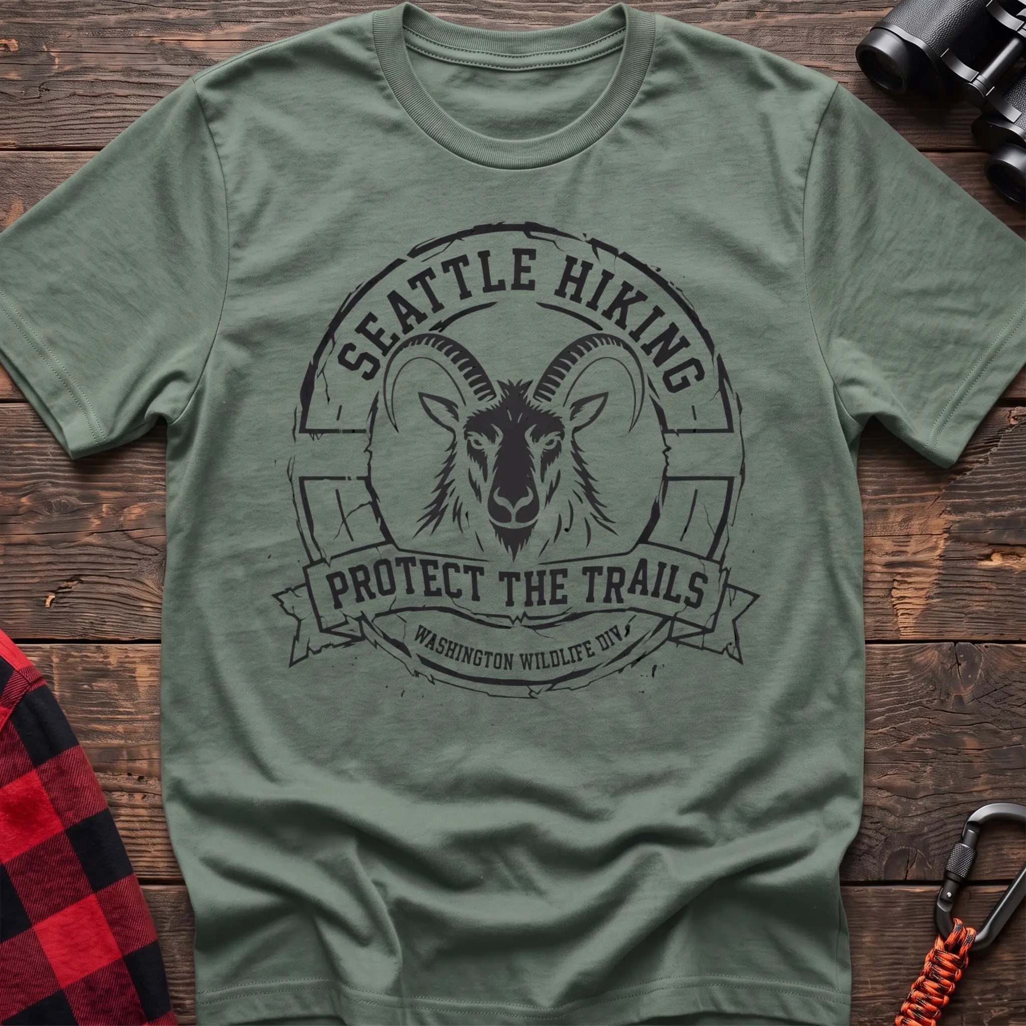 Sage cotton t-shirt with vintage Seattle Hiking graphic of a mountain goat in a circular badge, Protect the Trails Washington Wildlife Division design, flat lay on rustic wood with hiking gear
