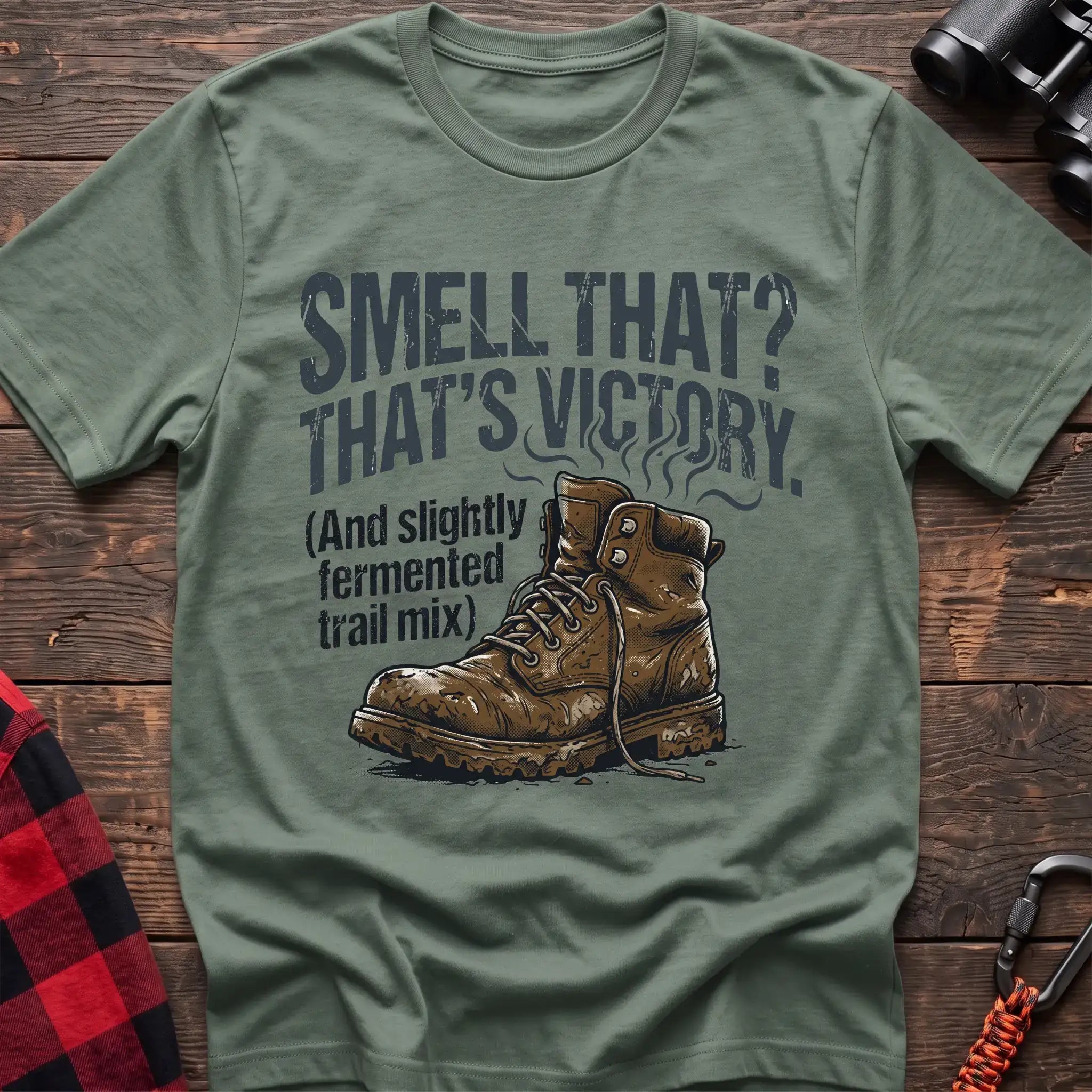 Sage t-shirt with an illustration of a dirty hiking boot with rising steam lines, and the text “SMELL THAT? THAT’S VICTORY. (And slightly fermented trail mix),” laid flat on a wooden surface
