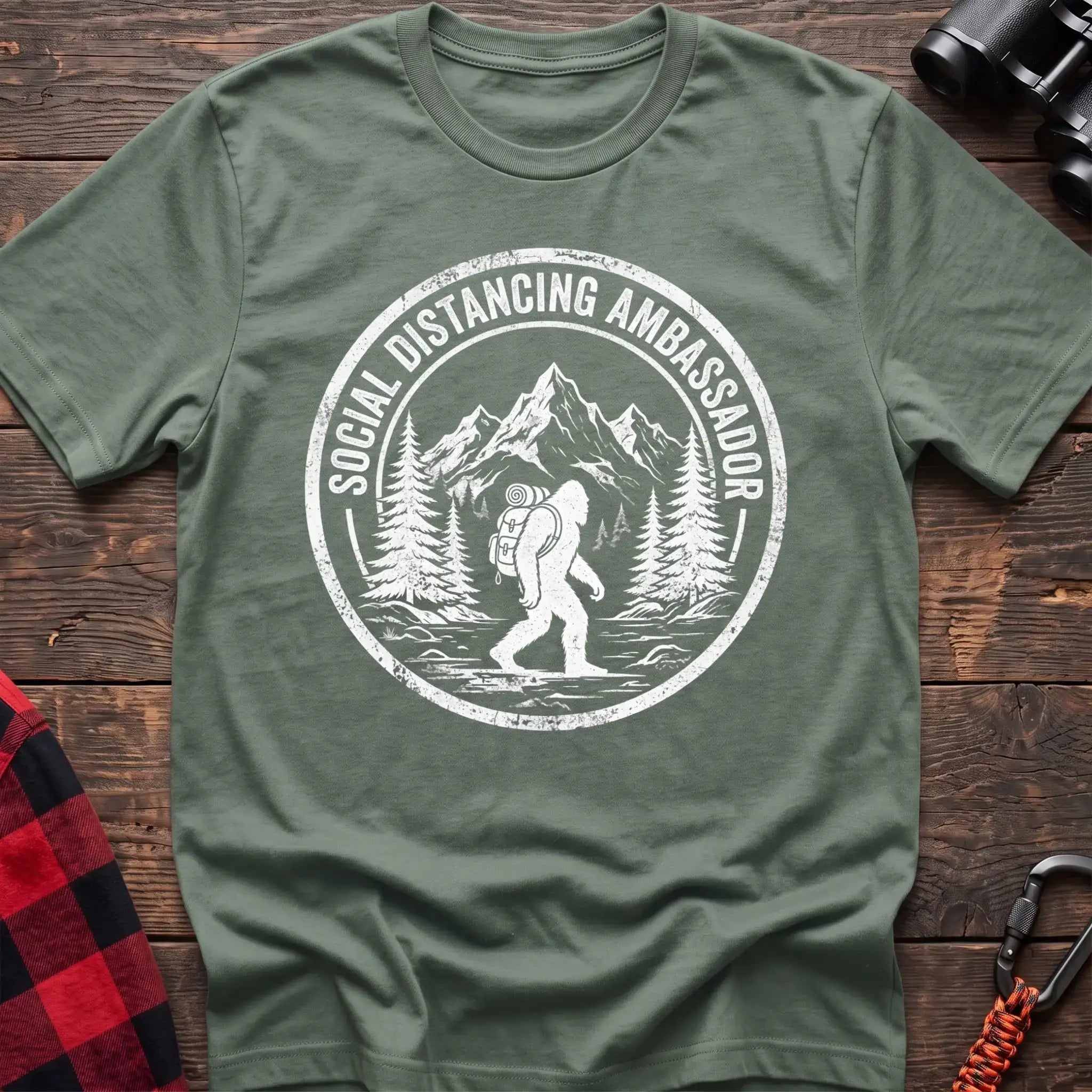 Sage t-shirt with white circular badge graphic of Bigfoot hiking with a backpack through mountains and the text Social Distancing Ambassador