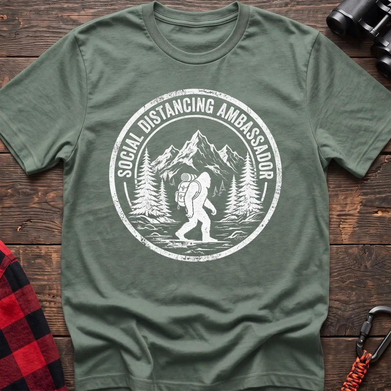 Sage t-shirt with white circular badge graphic of Bigfoot hiking with a backpack through mountains and the text Social Distancing Ambassador