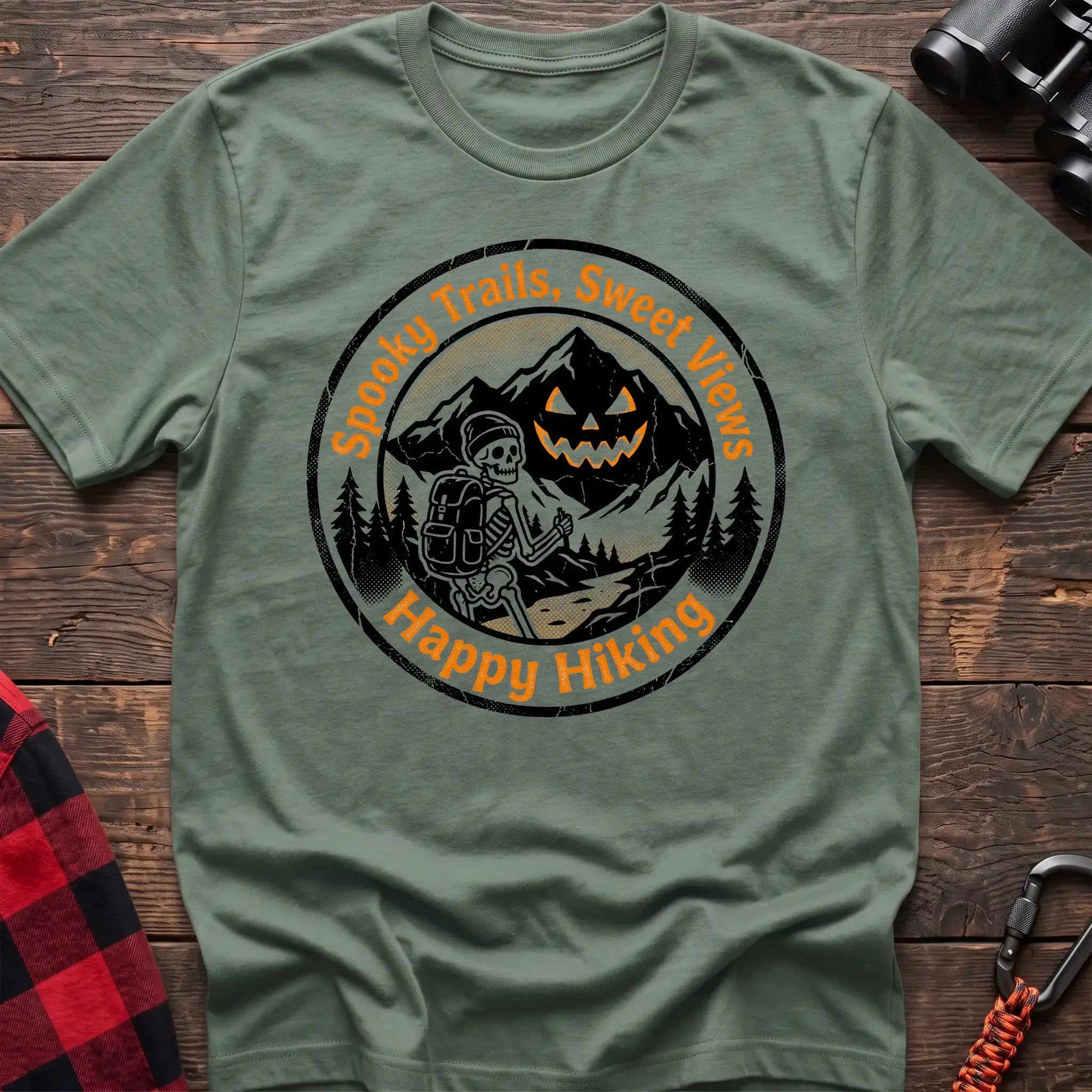 Sage green t-shirt with circular graphic of a skeleton hiker giving a thumbs up in front of a jack-o-lantern mountain, reading Spooky Trails Sweet Views Happy Hiking