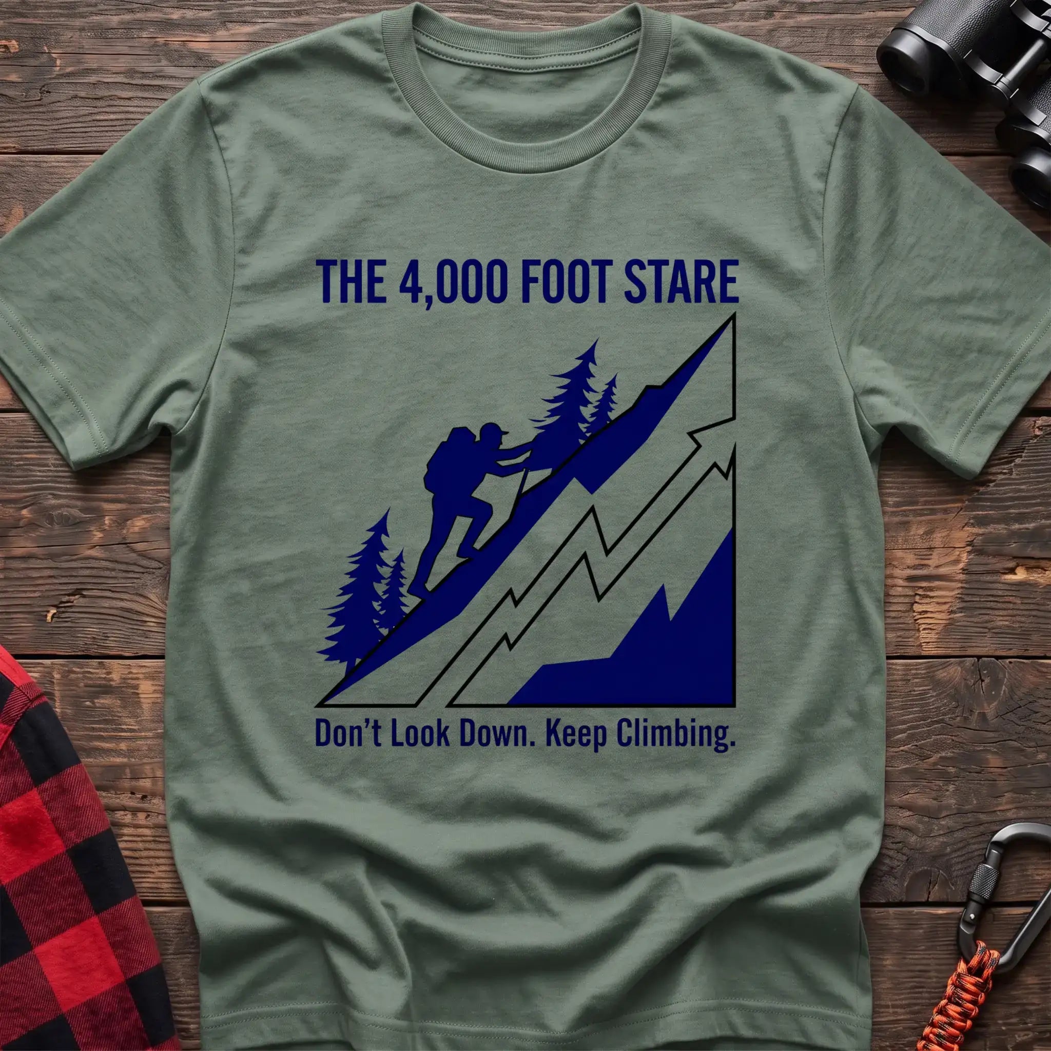 Sage t-shirt with a blue illustration of a hiker climbing a steep mountain, with the text “THE 4,000 FOOT STARE” and “Don’t Look Down. Keep Climbing,” laid flat on a wooden surface.