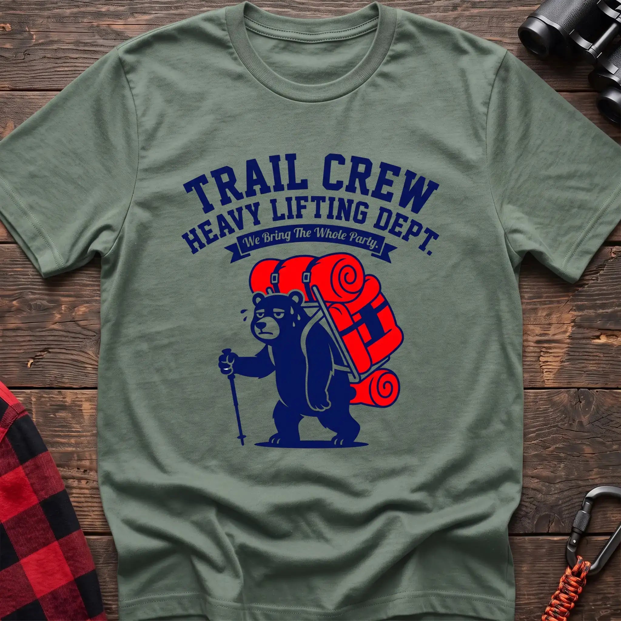 Sage t-shirt with a cartoon dark blue bear carrying a large red backpack and hiking pole, with the text “TRAIL CREW HEAVY LIFTING DEPT. We Bring The Whole Party,” laid flat on a wooden surface.