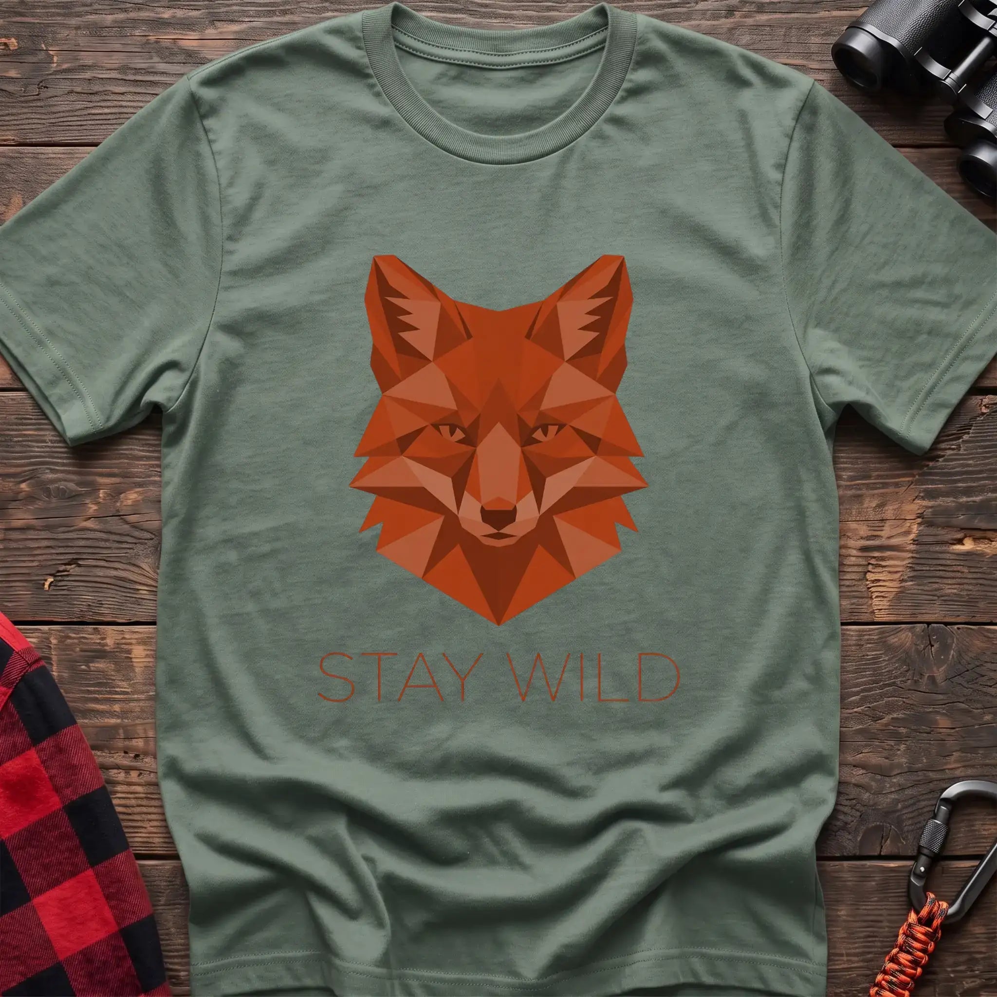 Sage t-shirt with a geometric low-poly orange fox head and the text “STAY WILD” printed below in matching orange, laid flat on a wooden surface.