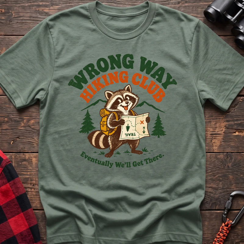 Sage cotton t-shirt with a raccoon holding a map graphic and Wrong Way Hiking Club text on rustic wood