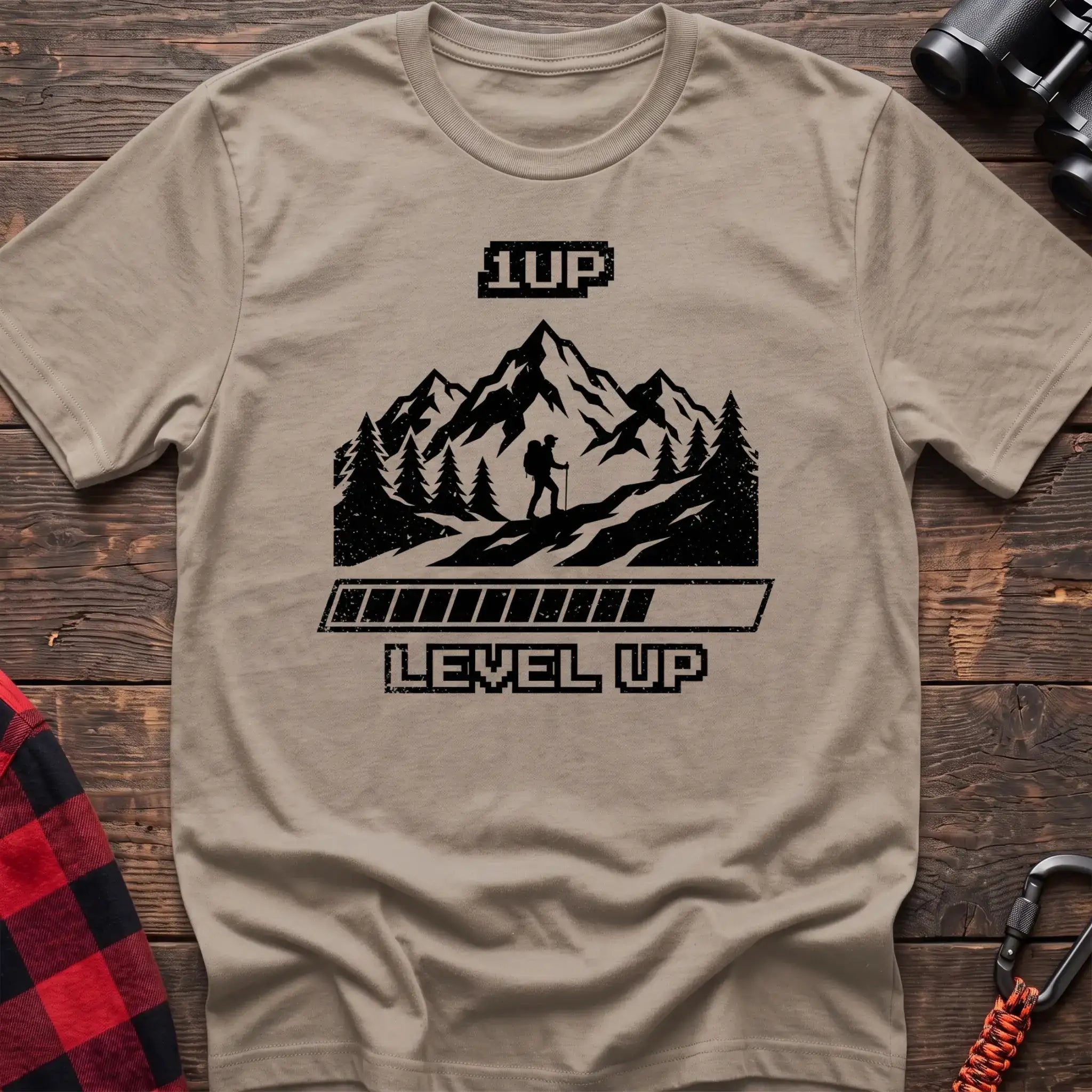 Sand t-shirt with black graphic of a hiker on a mountain trail under pixel-style 1UP text and a progress bar reading Level Up