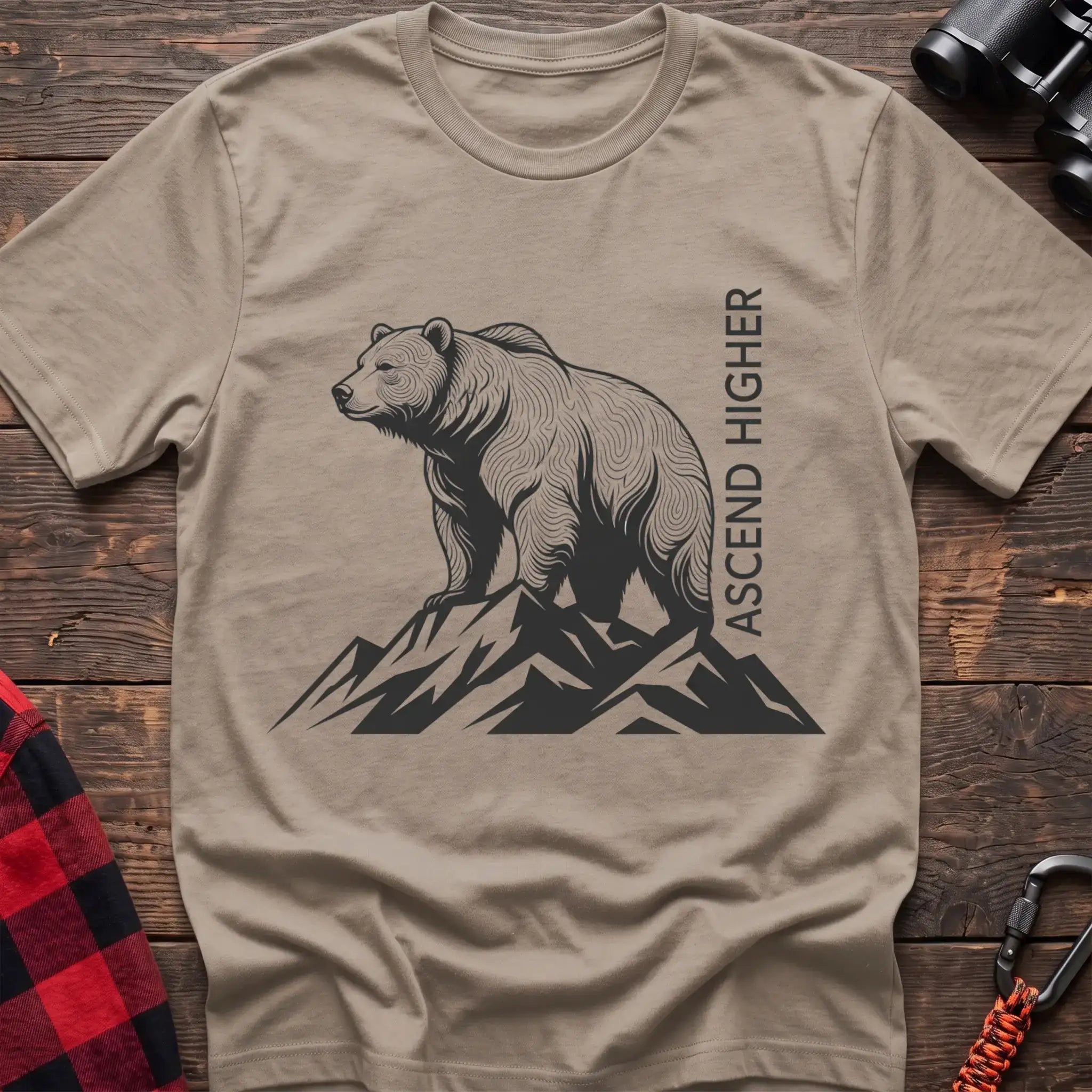 Sand t-shirt with a black illustration of a bear standing on mountain peaks, with the vertical text “ASCEND HIGHER” beside it, laid flat on a wooden surface.