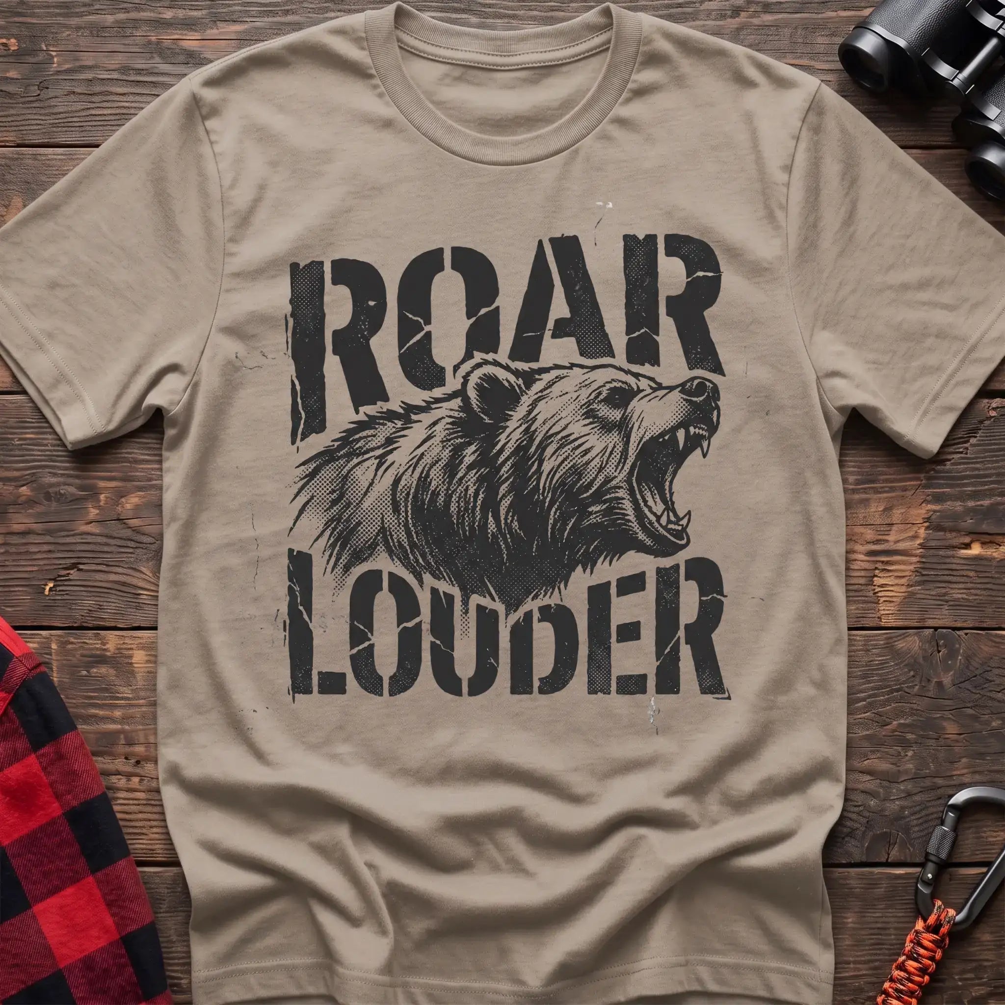 Sand t-shirt with a distressed black graphic of a roaring bear head between the bold text “ROAR” and “LOUDER,” laid flat on a wooden surface.