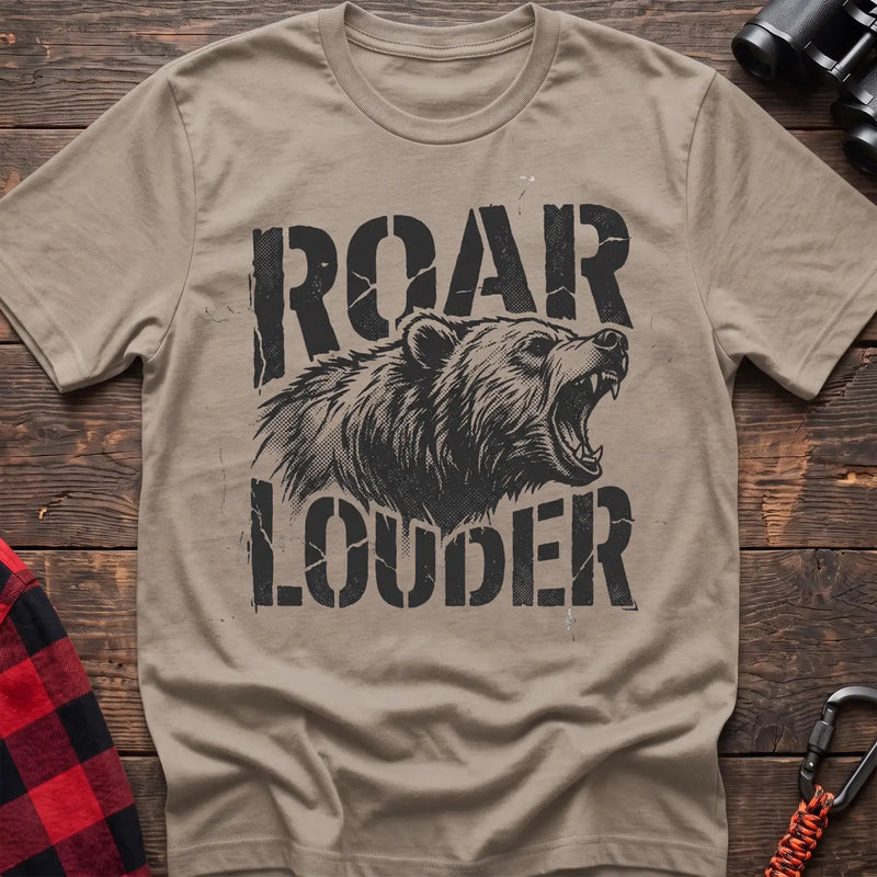 Sand t-shirt with a distressed black graphic of a roaring bear head between the bold text “ROAR” and “LOUDER,” laid flat on a wooden surface.