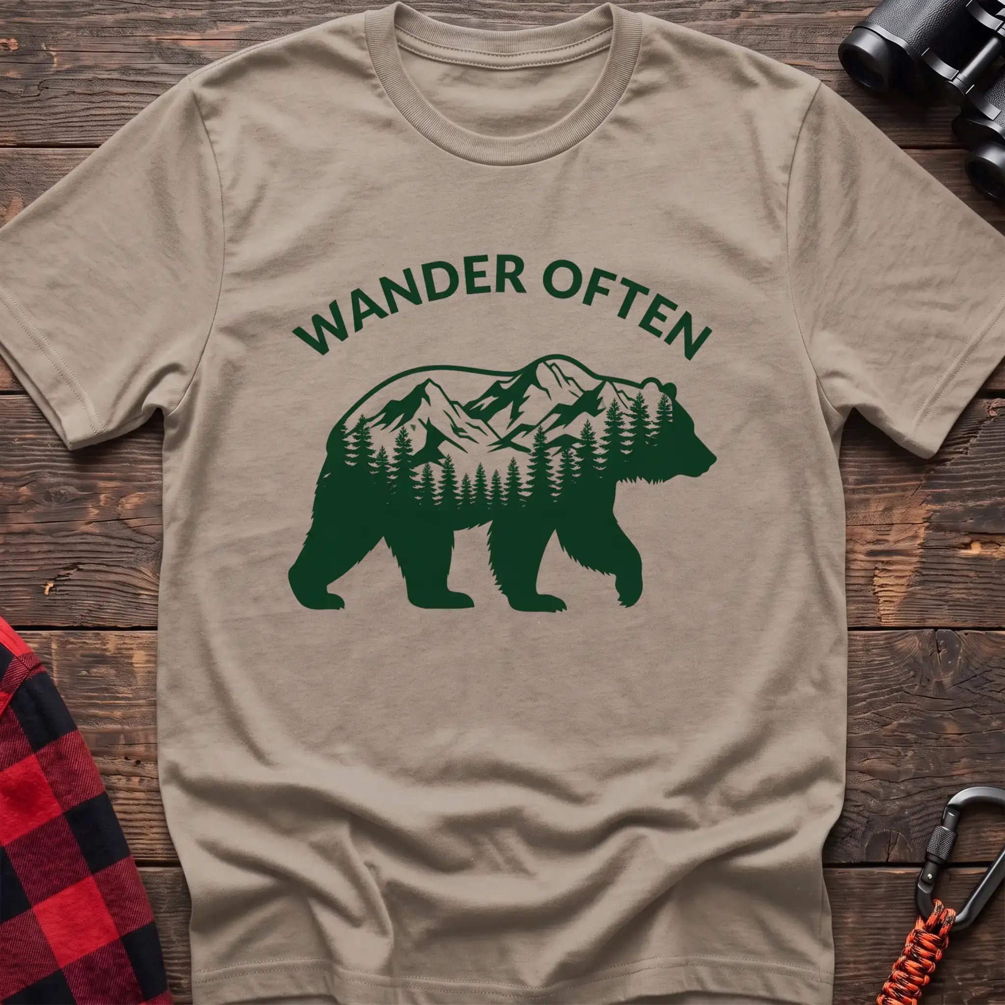 Sand t-shirt with a dark green silhouette of a walking bear containing a mountain and pine forest scene inside, with the arched text “WANDER OFTEN” above it, laid flat on a wooden surface.
