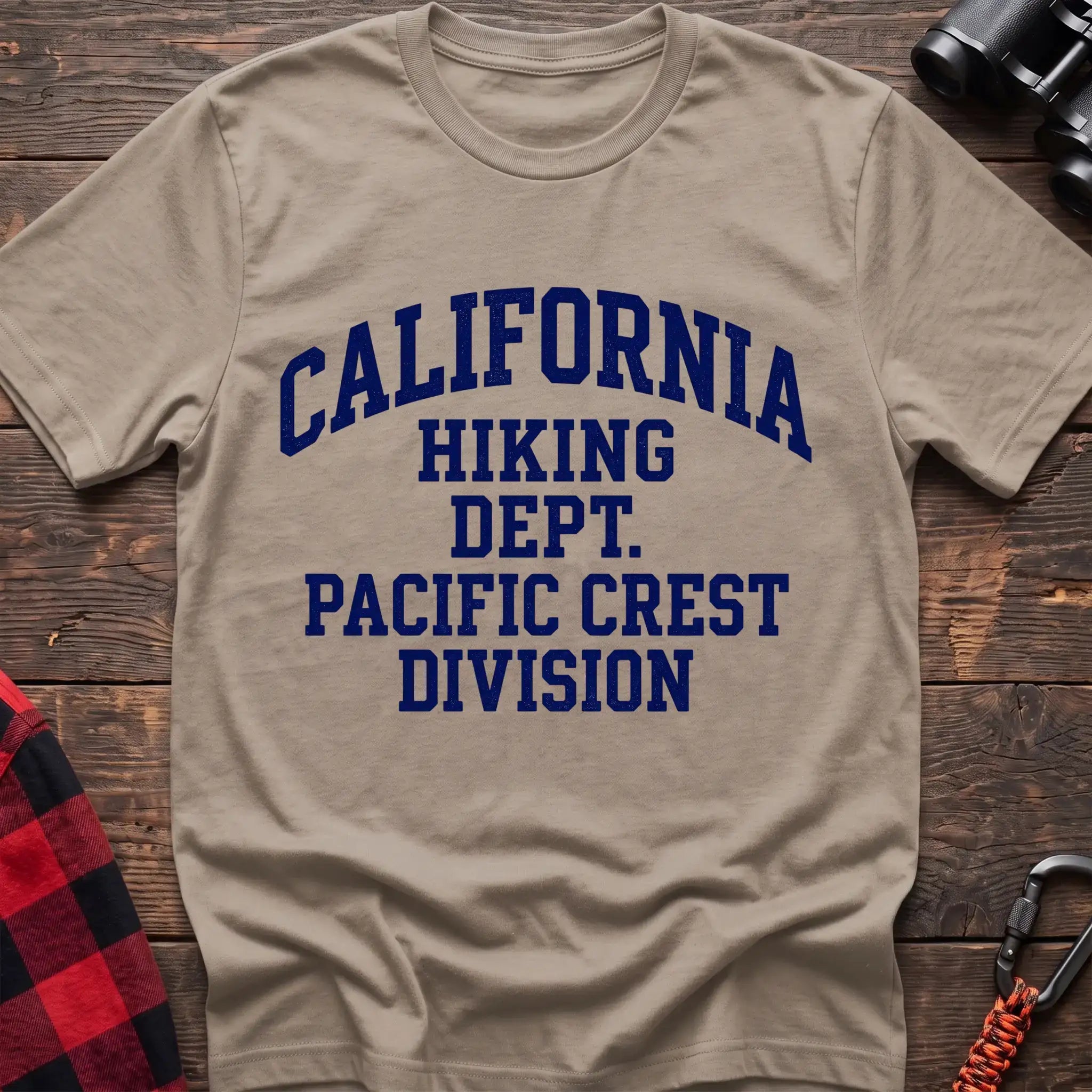 Sand t-shirt with distressed blue collegiate-style text reading “CALIFORNIA HIKING DEPT. PACIFIC CREST DIVISION,” laid flat on a wooden surface.