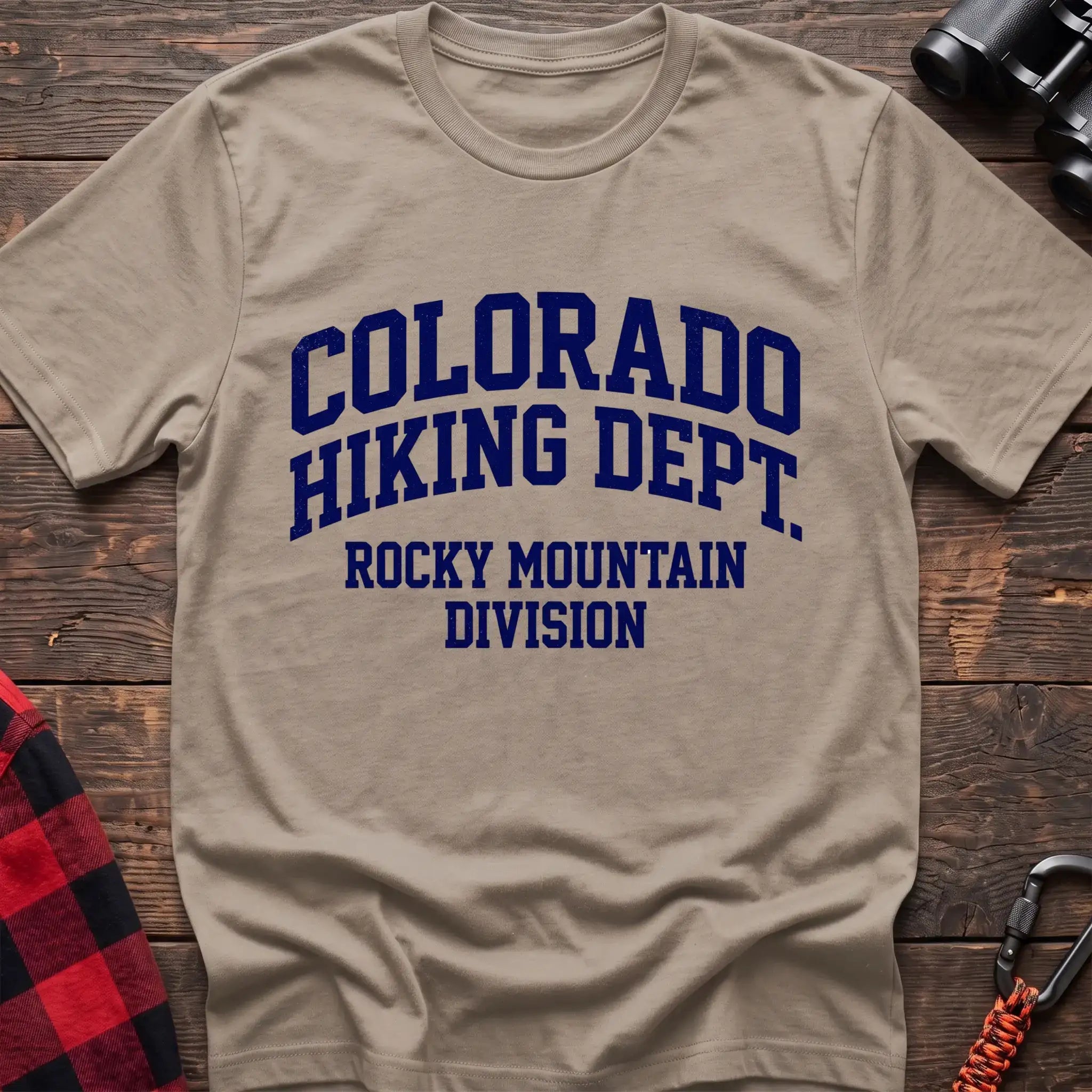Sand t-shirt with distressed dark blue collegiate-style text reading “COLORADO HIKING DEPT. ROCKY MOUNTAIN DIVISION,” laid flat on a wooden surface.