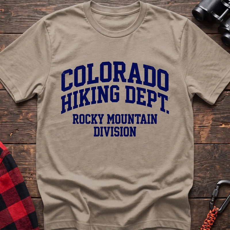 Sand t-shirt with distressed dark blue collegiate-style text reading “COLORADO HIKING DEPT. ROCKY MOUNTAIN DIVISION,” laid flat on a wooden surface.