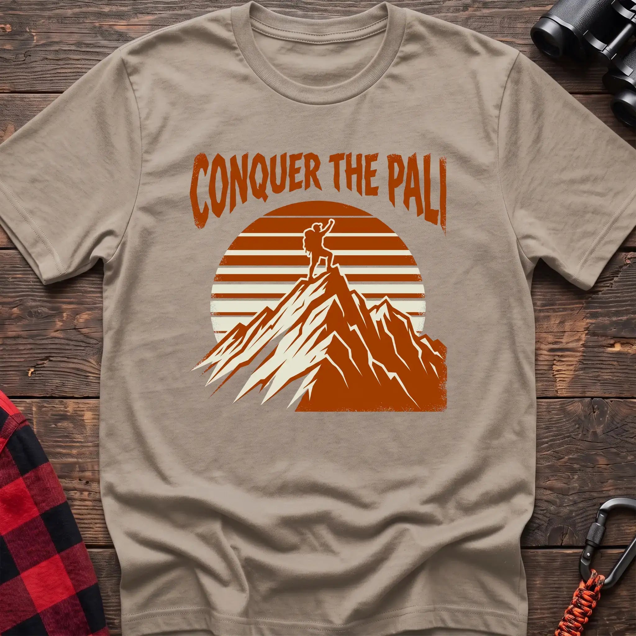 Sand t-shirt with orange text reading Conquer the Pali above a hiker standing on a mountain peak with a retro sun graphic