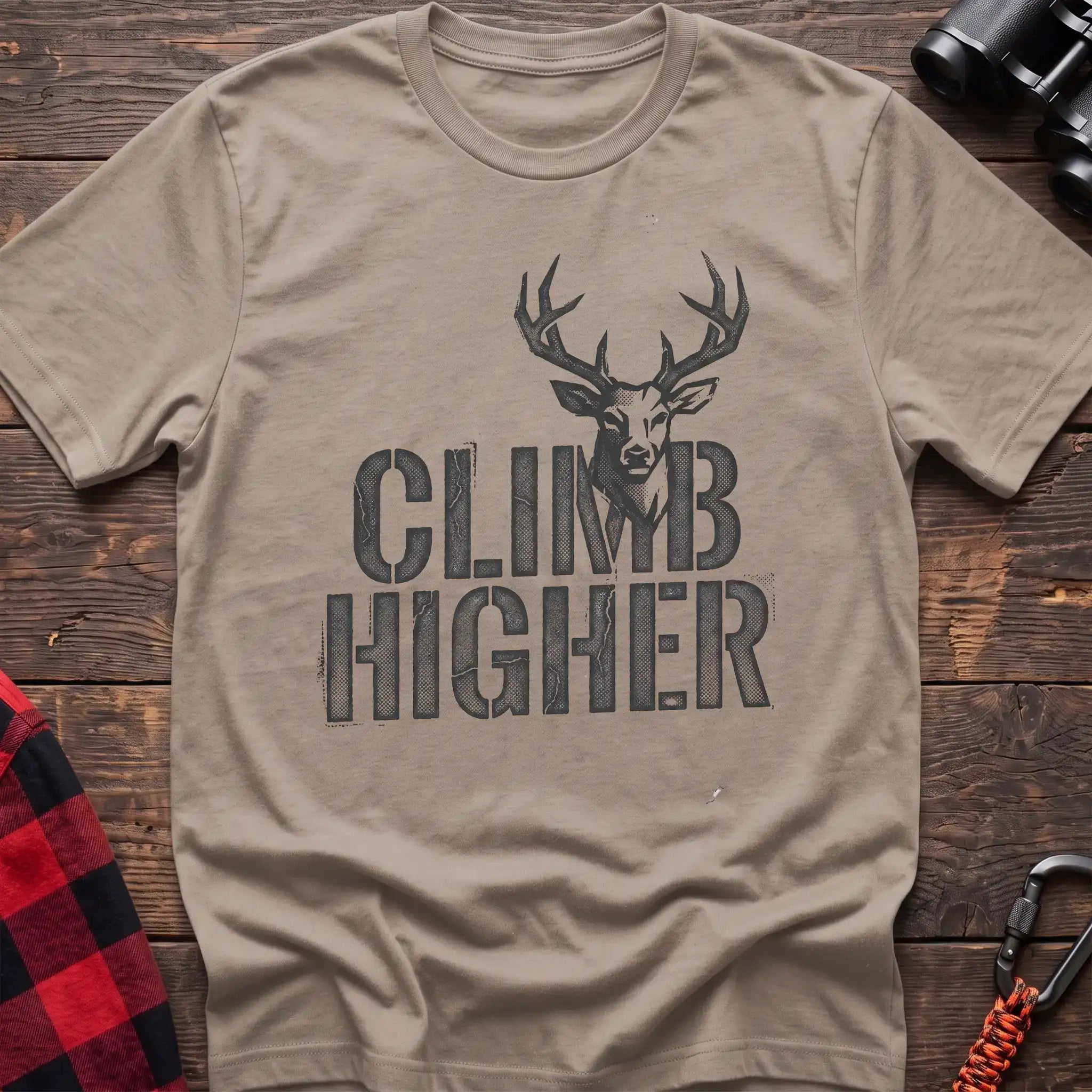 Sand t-shirt with a distressed dark graphic of a deer’s head and antlers rising from the bold text “CLIMB HIGHER,” laid flat on a wooden surface.