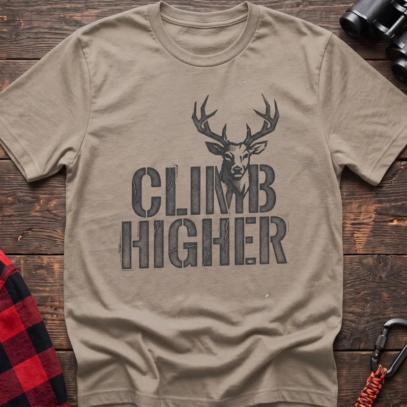 Sand t-shirt with a distressed dark graphic of a deer’s head and antlers rising from the bold text “CLIMB HIGHER,” laid flat on a wooden surface.