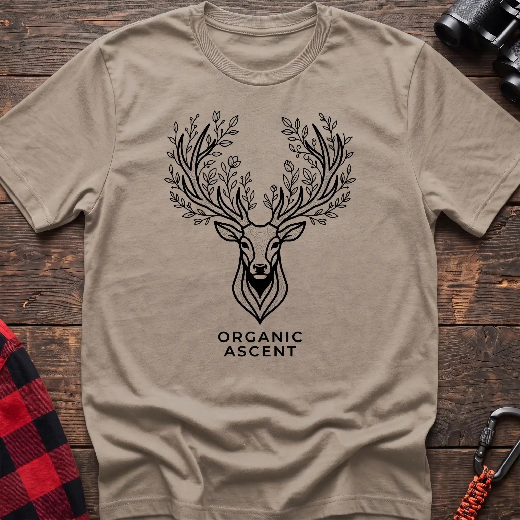 Sand t-shirt with a black line drawing of a deer’s head whose antlers are formed from leafy branches and flowers, with the text “ORGANIC ASCENT” printed below, laid flat on a wooden surface.