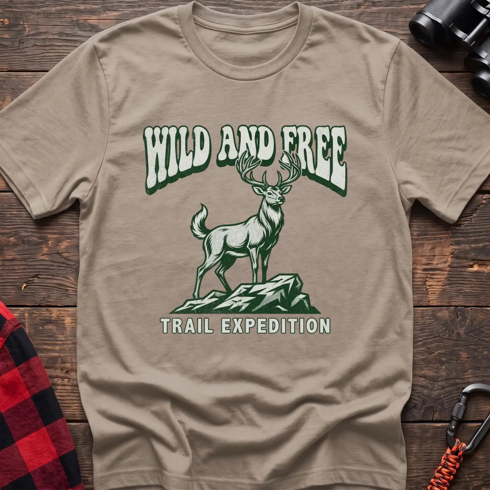 Sand t-shirt with a rustic graphic of a deer standing on a rocky outcrop, with the arched text “WILD AND FREE” above and “TRAIL EXPEDITION” below, laid flat on a wooden surface.