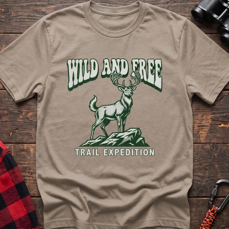 Sand t-shirt with a rustic graphic of a deer standing on a rocky outcrop, with the arched text “WILD AND FREE” above and “TRAIL EXPEDITION” below, laid flat on a wooden surface.