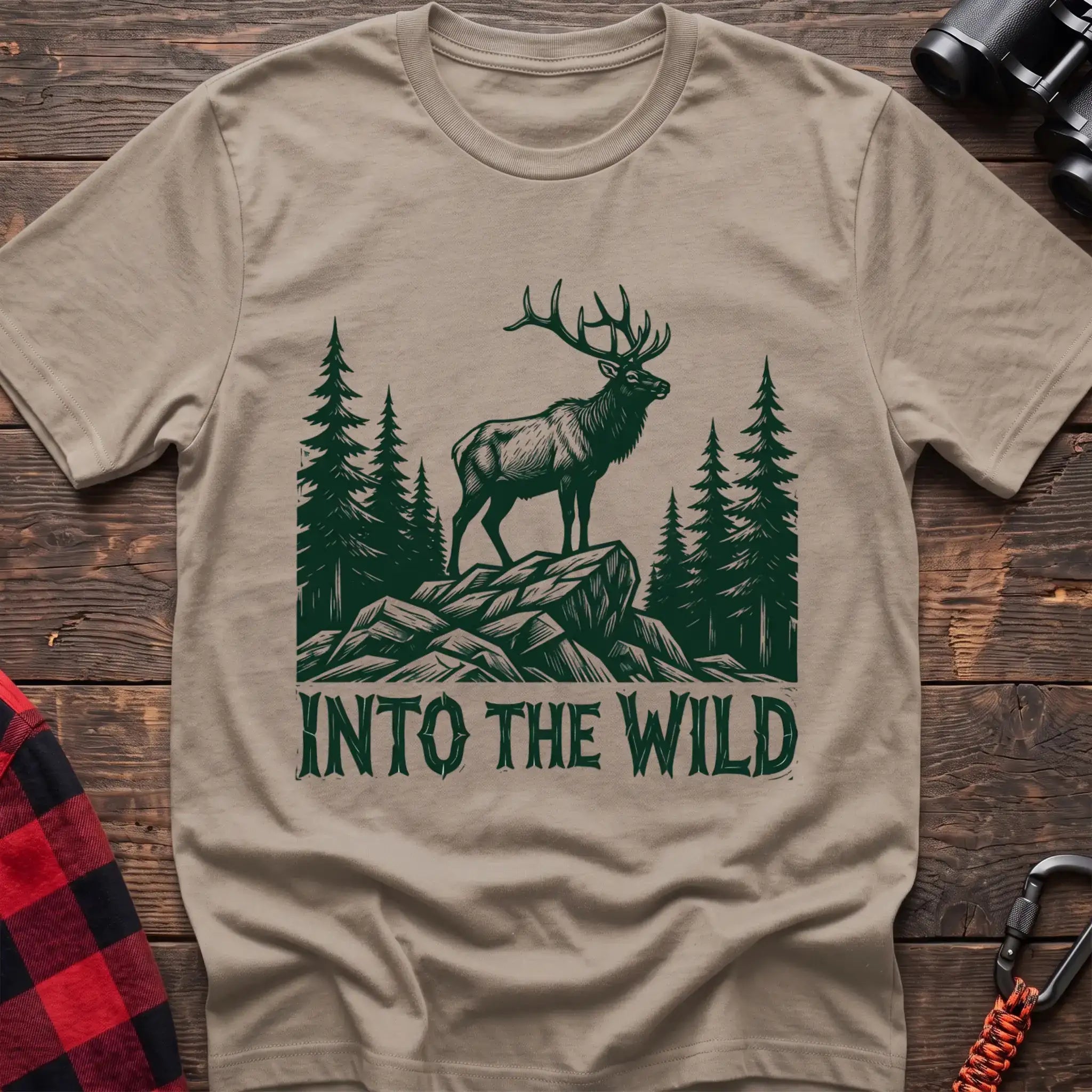 Sand t-shirt with a dark green illustration of an elk standing on a rocky outcrop among pine trees, with the text “INTO THE WILD” printed below, laid flat on a wooden surface.