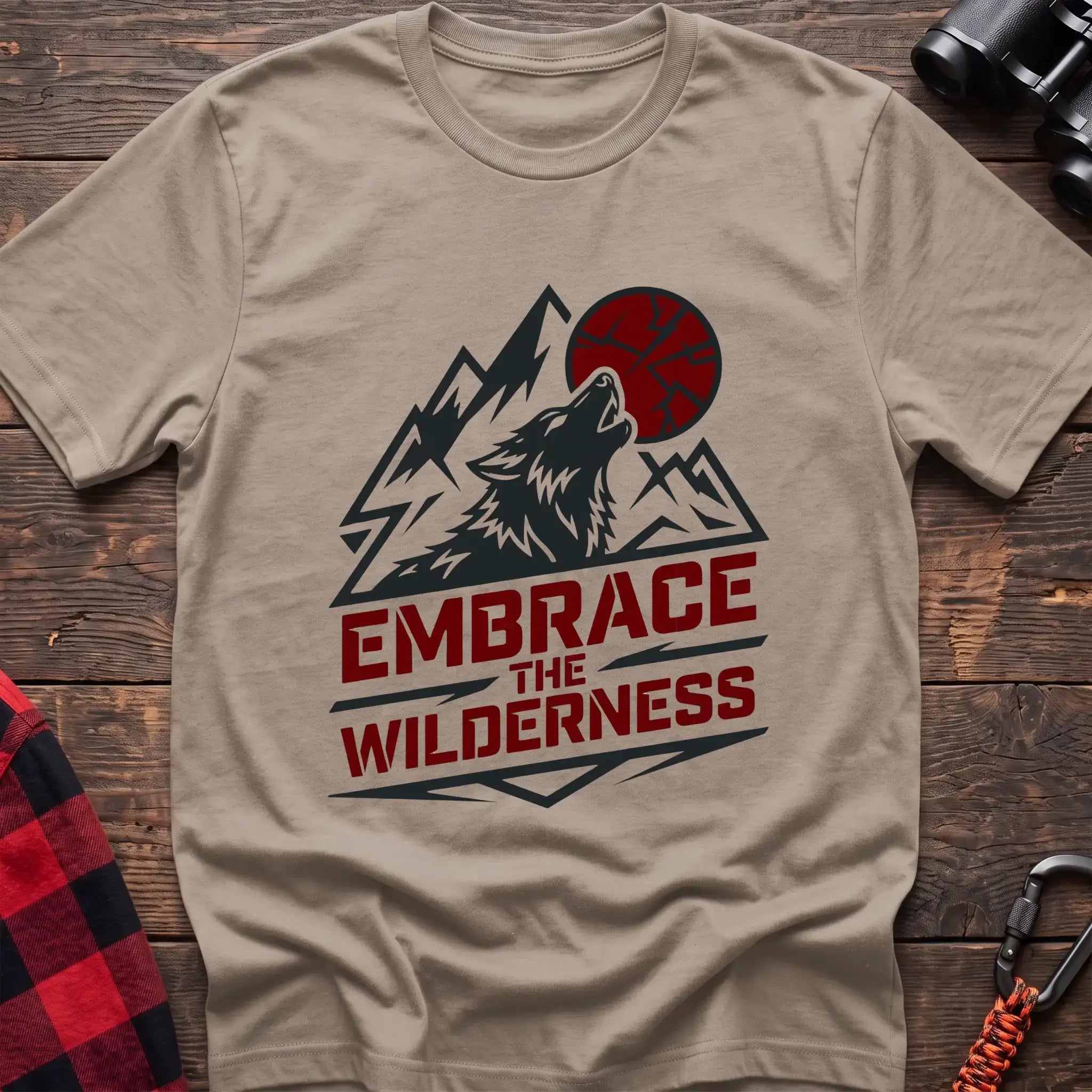 Sand t-shirt with a dark graphic of a howling wolf against mountain peaks and a red sun, with the bold red text “EMBRACE THE WILDERNESS” below, laid flat on a wooden surface.