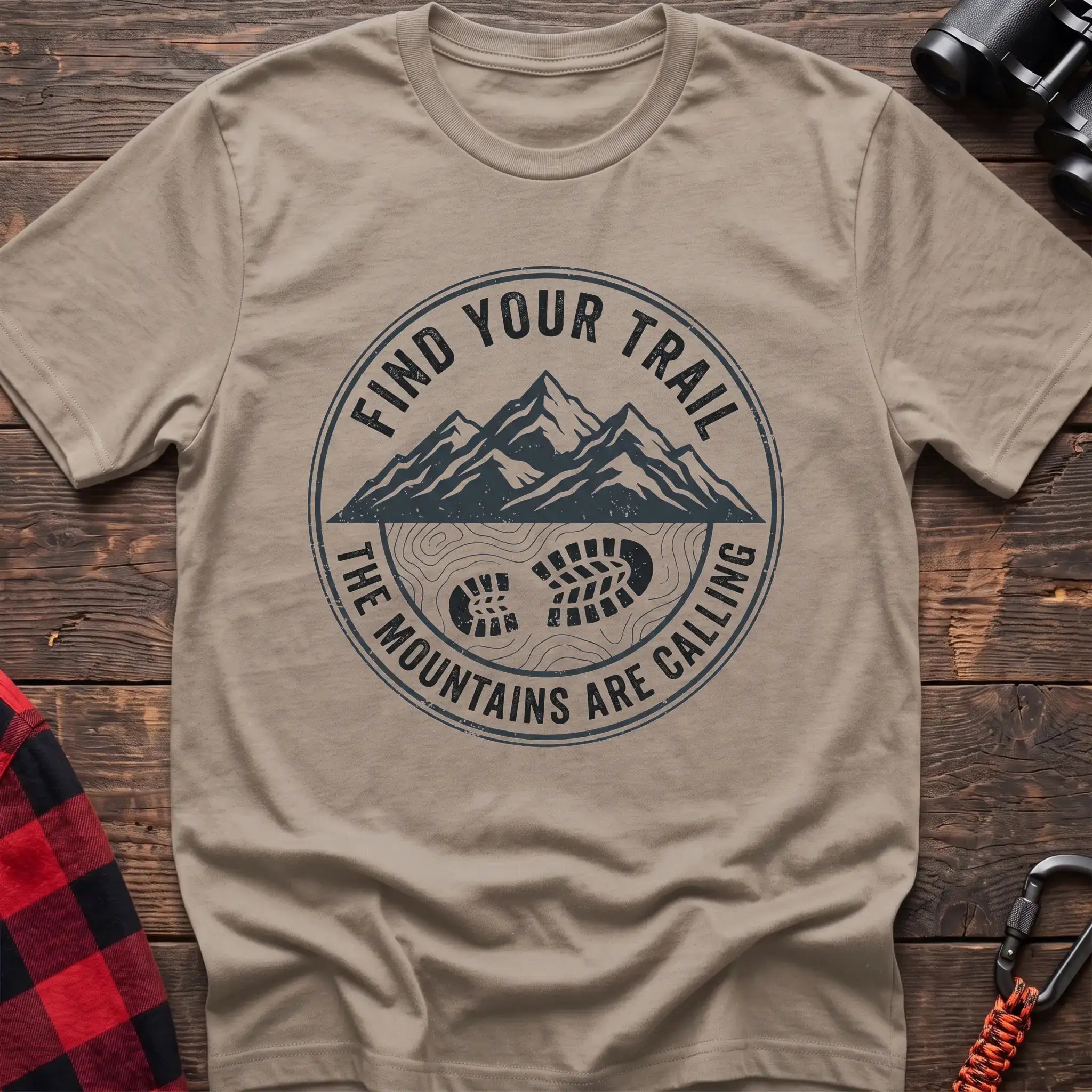 Sand t-shirt with a circular dark gray badge design featuring mountain peaks above a hiking boot print on topographic lines, with the text “FIND YOUR TRAIL” and “THE MOUNTAINS ARE CALLING,” laid flat on a wooden surface.