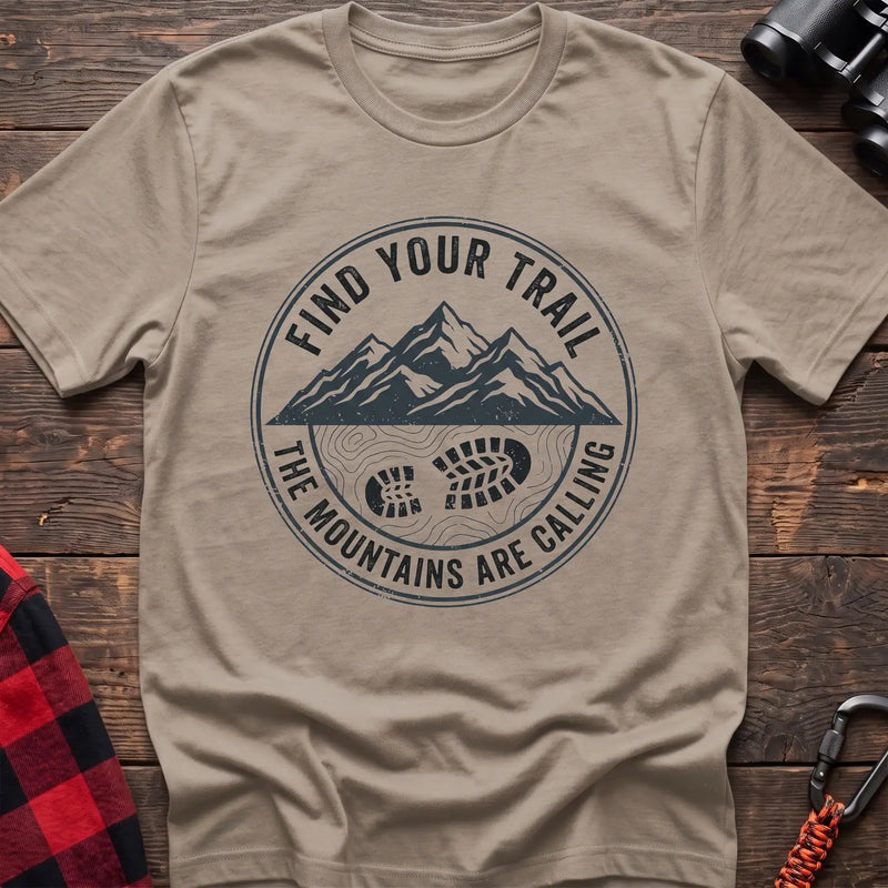 Sand t-shirt with a circular dark gray badge design featuring mountain peaks above a hiking boot print on topographic lines, with the text “FIND YOUR TRAIL” and “THE MOUNTAINS ARE CALLING,” laid flat on a wooden surface.
