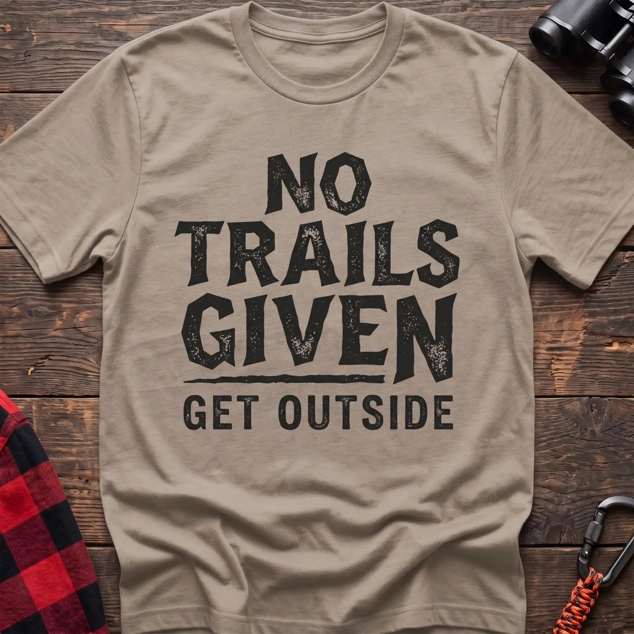Sand t-shirt with distressed black text reading “NO TRAILS GIVEN” and “GET OUTSIDE” below it, laid flat on a wooden surface.