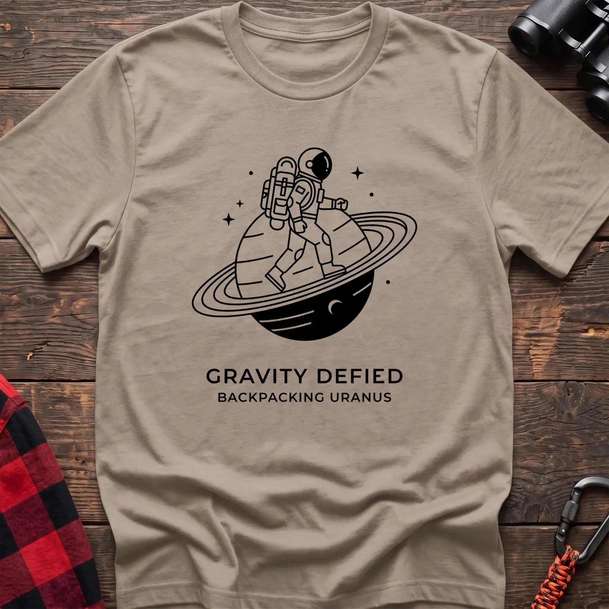 Sand crew-neck t-shirt with black line-art graphic of an astronaut backpacking across a ringed planet surrounded by stars, with the text “GRAVITY DEFIED / BACKPACKING URANUS” printed below, laid flat on a wooden surface.