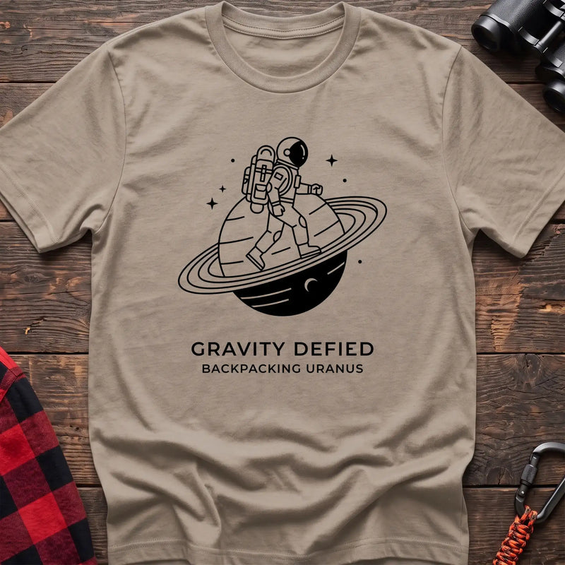 Sand crew-neck t-shirt with black line-art graphic of an astronaut backpacking across a ringed planet surrounded by stars, with the text “GRAVITY DEFIED / BACKPACKING URANUS” printed below, laid flat on a wooden surface.