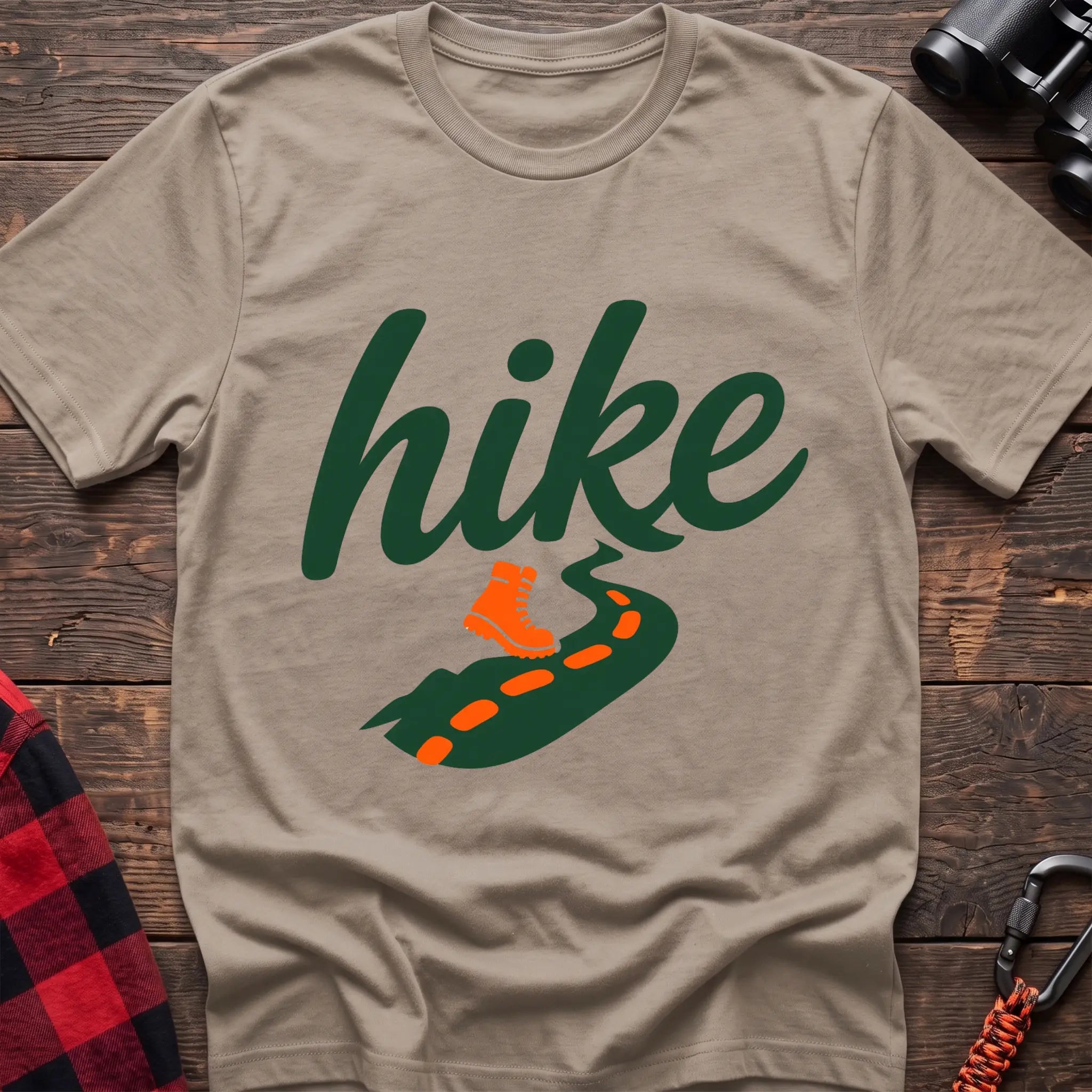 Sand cotton t-shirt with green cursive hike script, orange hiking boot, and footprint trail graphic, flat lay on rustic wood with hiking gear
