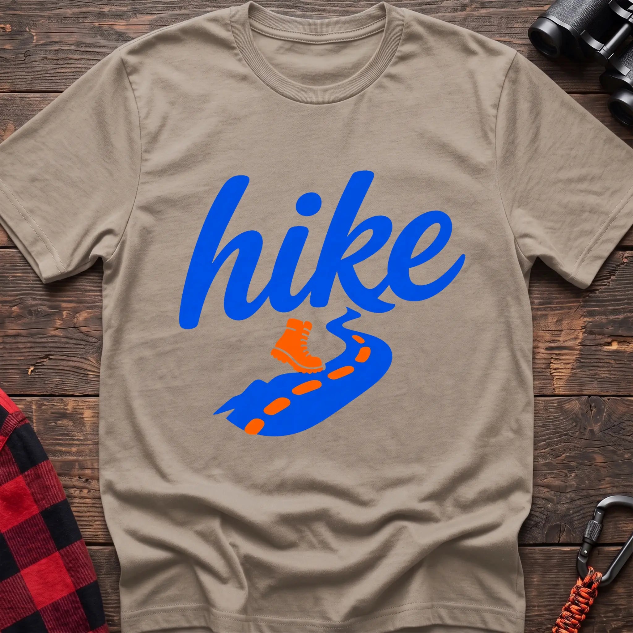Sand cotton t-shirt with royal blue cursive hike script, orange hiking boot, and blue trail with orange footprints, flat lay on rustic wood with hiking gear