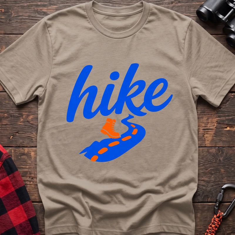 Sand cotton t-shirt with royal blue cursive hike script, orange hiking boot, and blue trail with orange footprints, flat lay on rustic wood with hiking gear