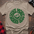 Sand t-shirt with a circular green design featuring mountains and pine trees in the center, surrounded by the text “HIKE MORE WORRY LESS,” laid flat on a wooden surface.