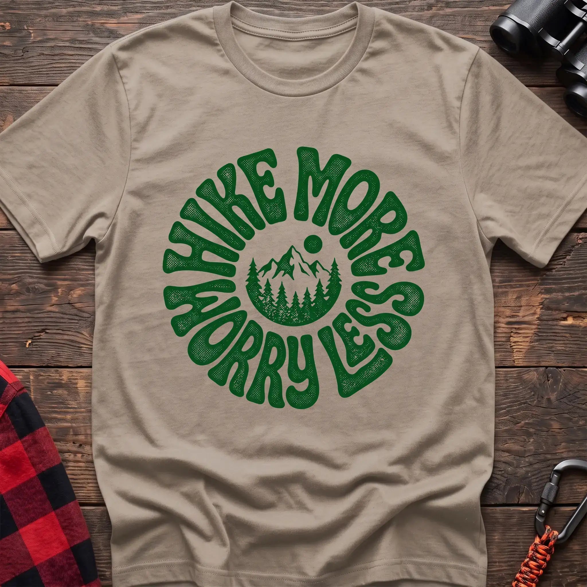 Sand t-shirt with a circular green design featuring mountains and pine trees in the center, surrounded by the text “HIKE MORE WORRY LESS,” laid flat on a wooden surface.