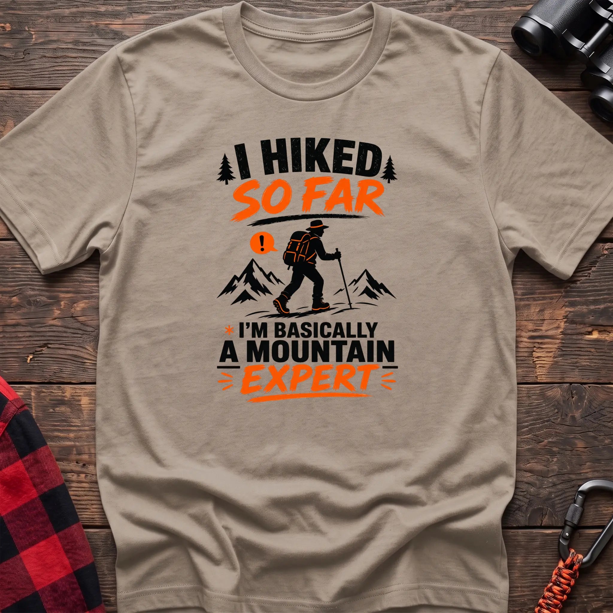 Sand cotton t-shirt with I Hiked So Far I'm Basically a Mountain Expert graphic of a hiker with backpack and trekking pole, flat lay on rustic wood with hiking gear