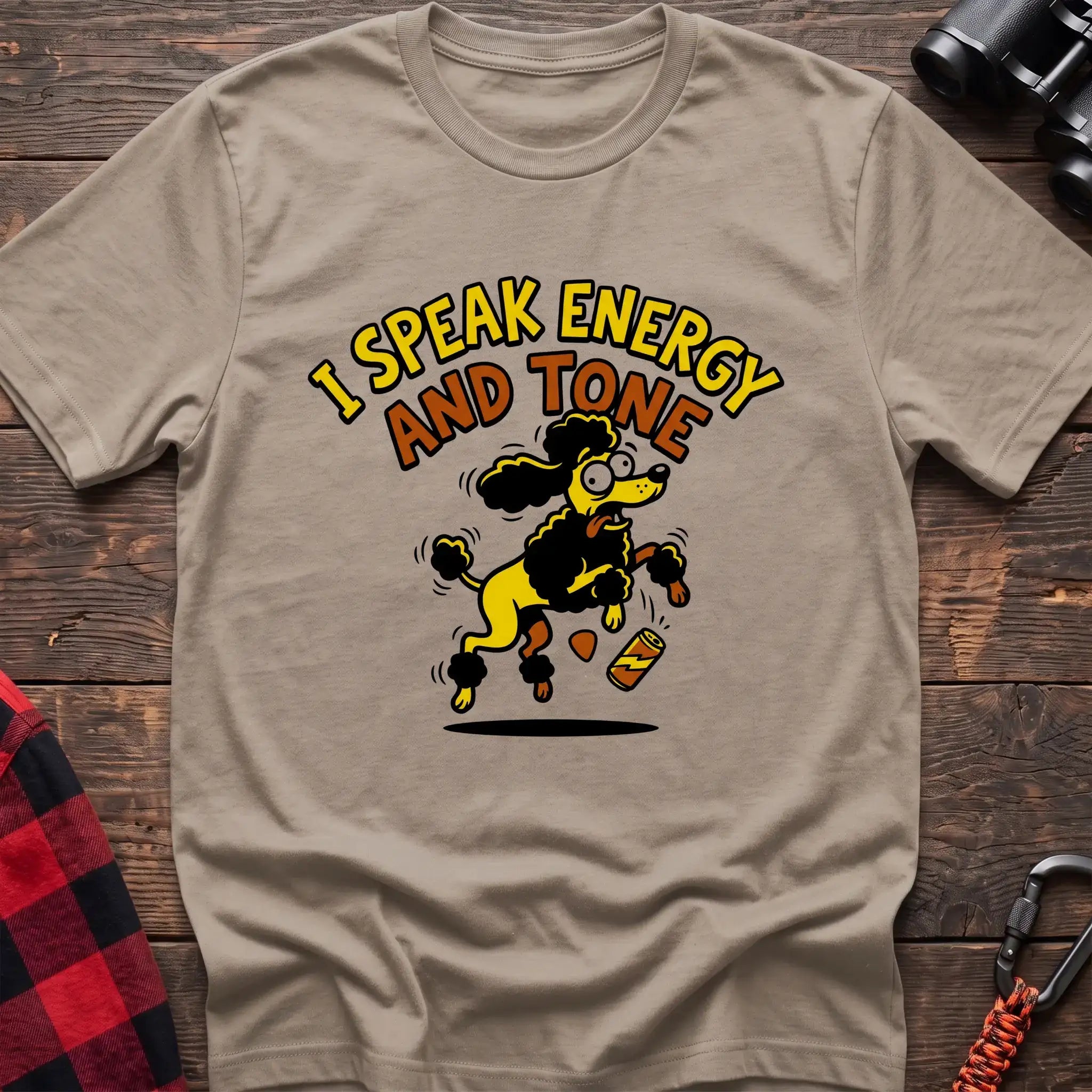 Sand t-shirt with a cartoon yellow and black poodle jumping excitedly next to a falling energy drink can, with the arched text “I SPEAK ENERGY AND TONE,” laid flat on a wooden surface.