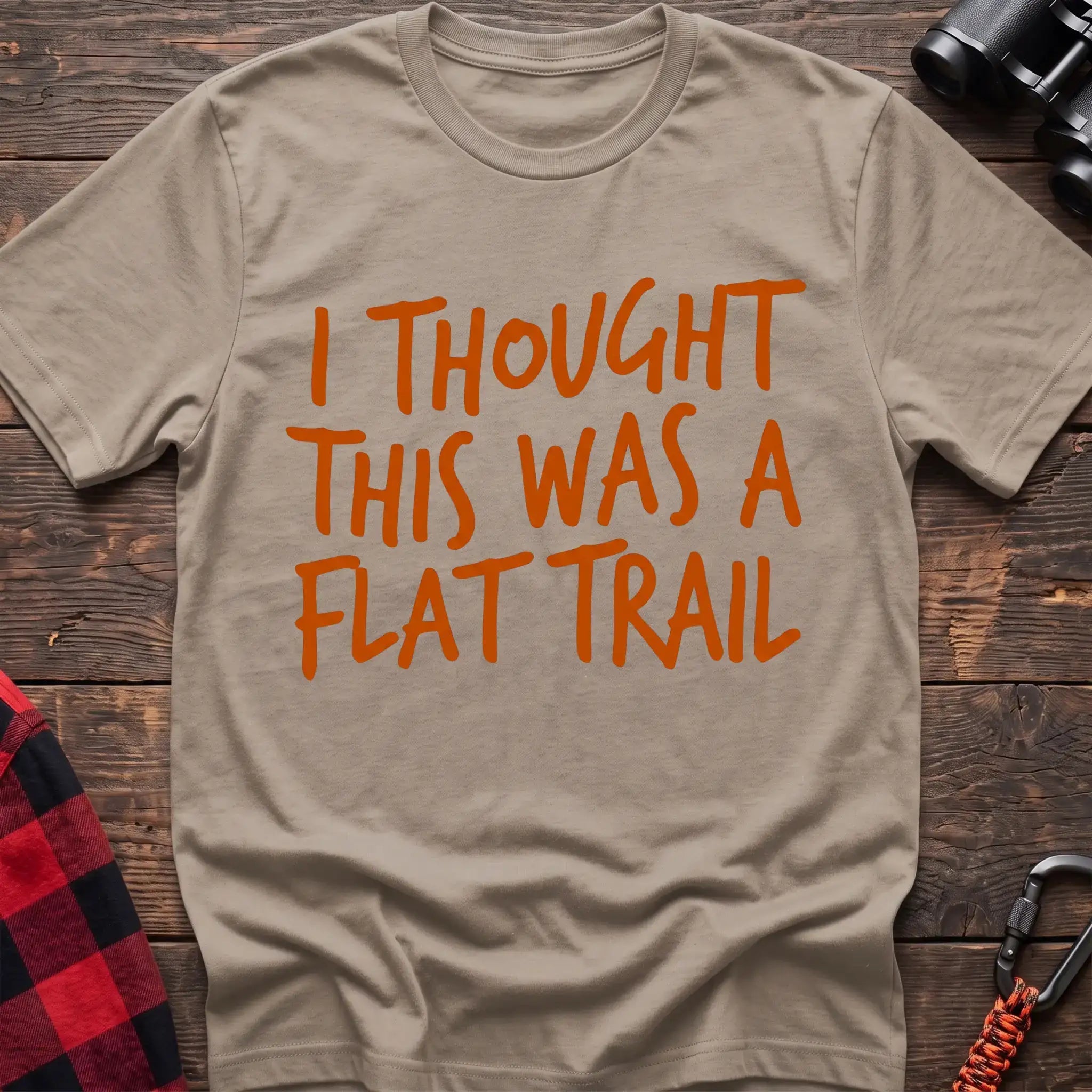 Sand t-shirt with bold orange handwritten-style text reading “I THOUGHT THIS WAS A FLAT TRAIL,” laid flat on a wooden surface.