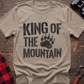 Sand t-shirt with distressed black text reading “KING OF THE MOUNTAIN” and a large bear paw print integrated into the design, laid flat on a wooden surface.