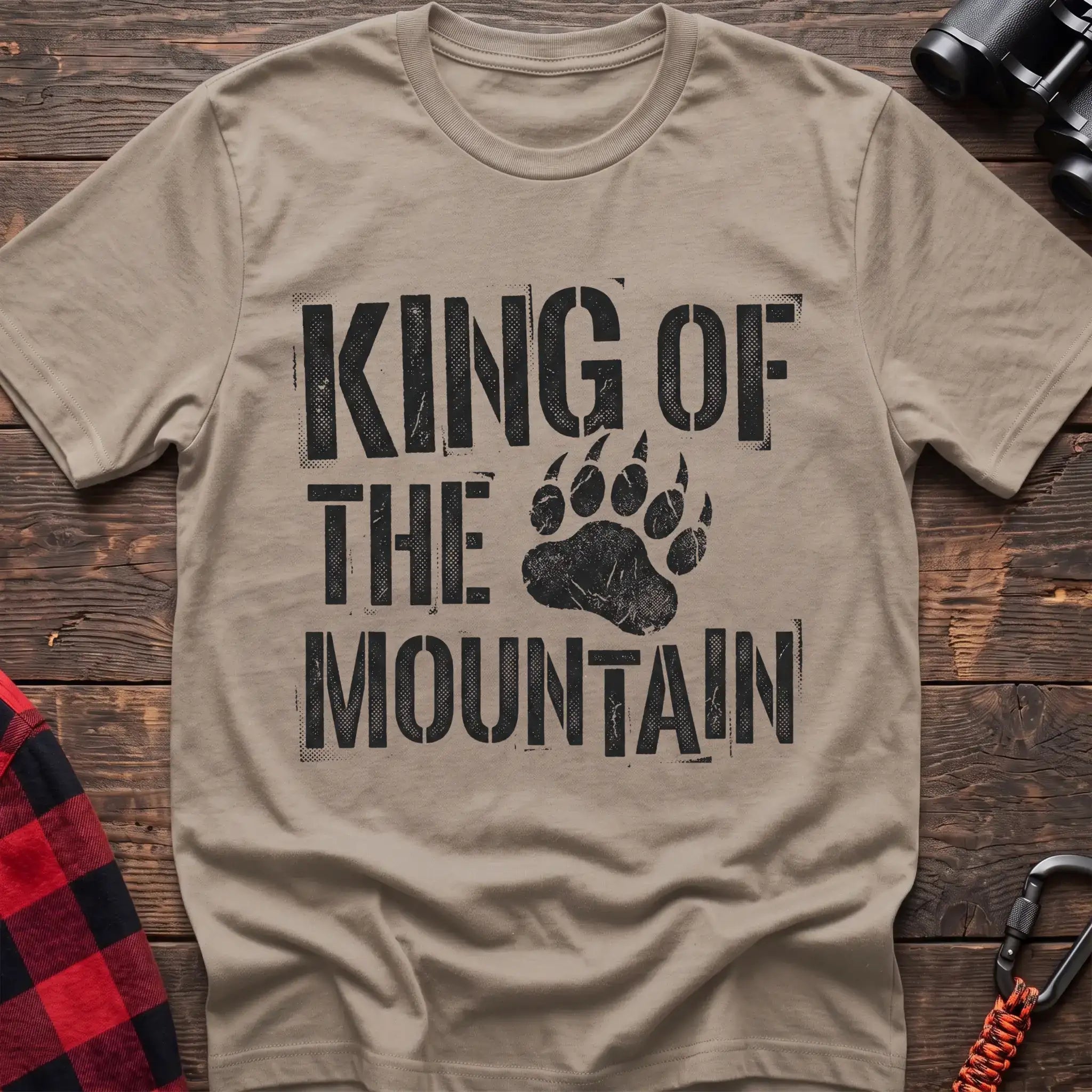 Sand t-shirt with distressed black text reading “KING OF THE MOUNTAIN” and a large bear paw print integrated into the design, laid flat on a wooden surface.