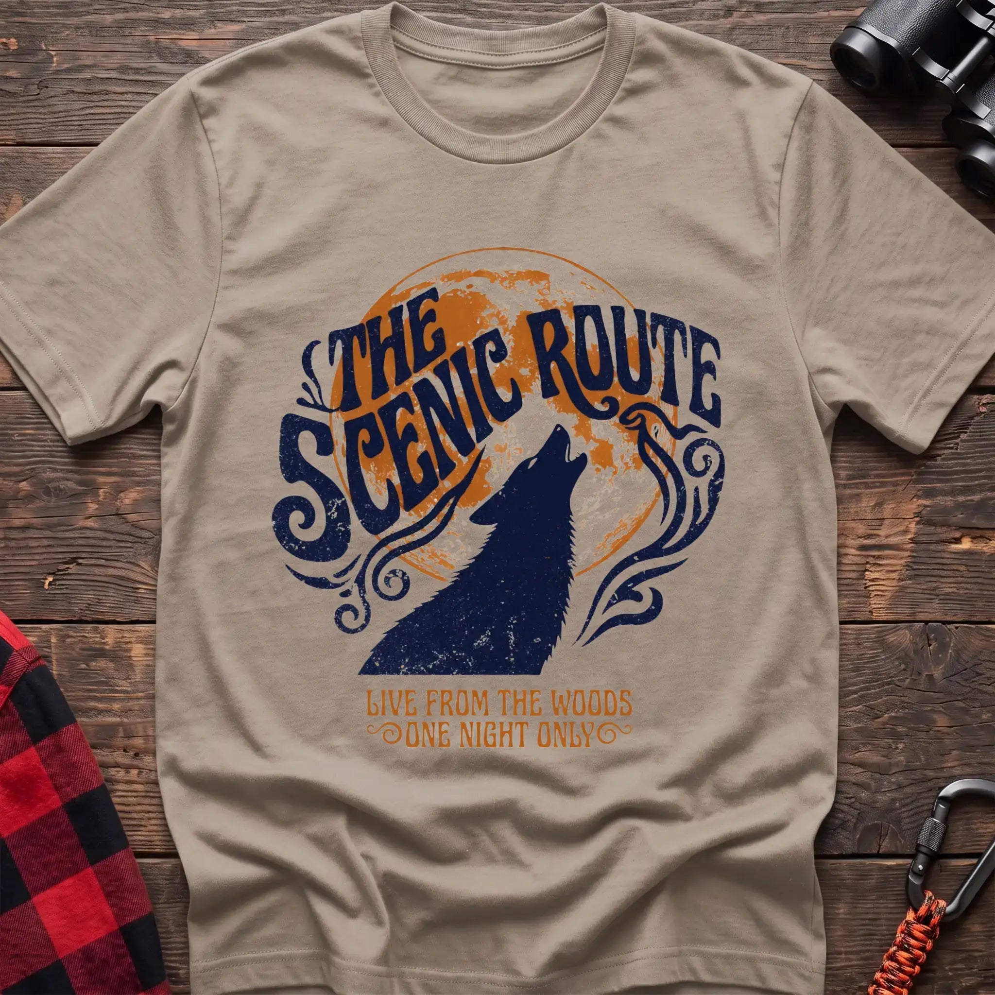 Sand t-shirt with a dark blue howling wolf silhouette against an orange circle, with the arched text “THE SCENIC ROUTE” above and “LIVE FROM THE WOODS / ONE NIGHT ONLY” below, laid flat on a wooden surface.