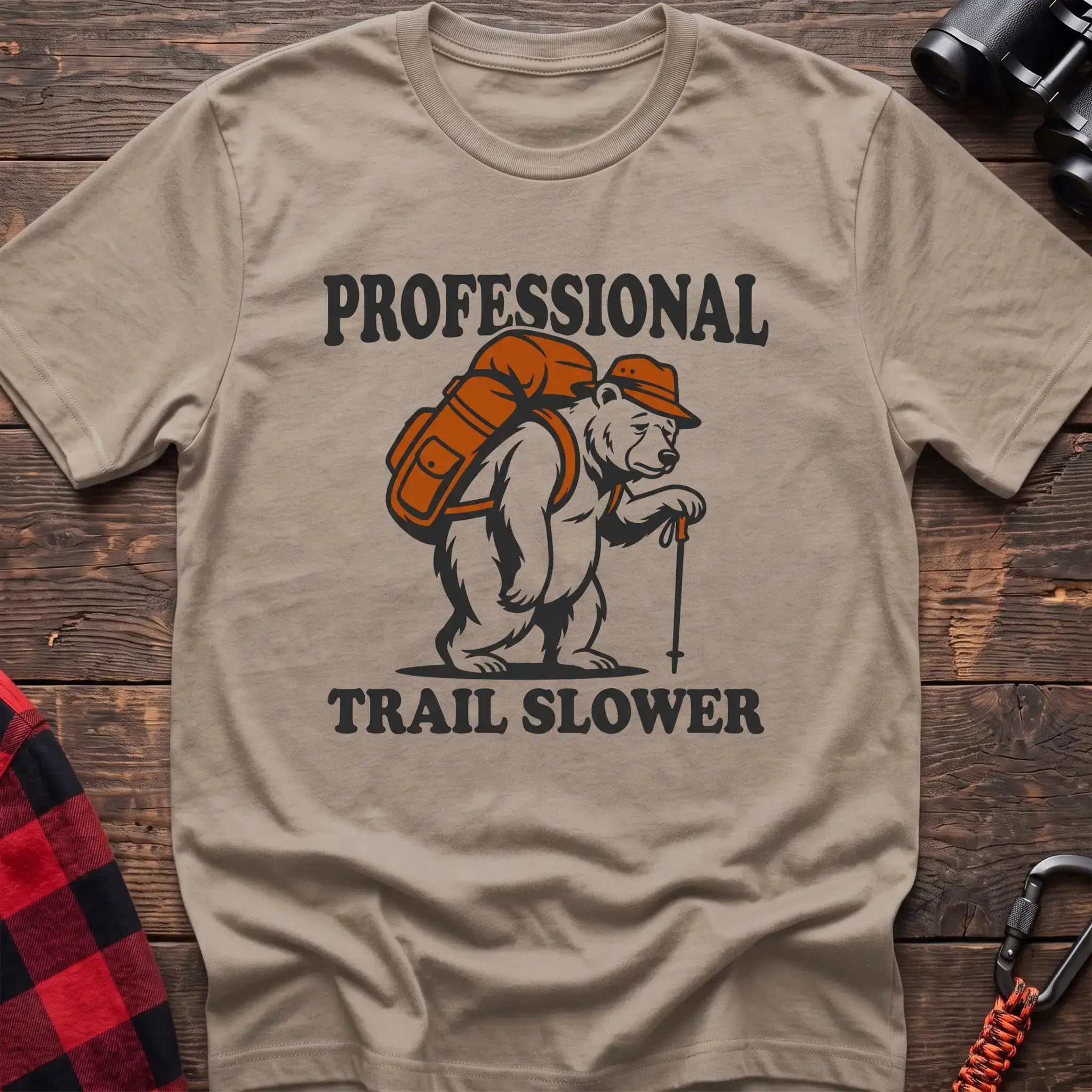 Sand t-shirt with a cartoon polar bear wearing a backpack and hat while holding a hiking stick, with the text “PROFESSIONAL TRAIL SLOWER,” laid flat on a wooden surface.