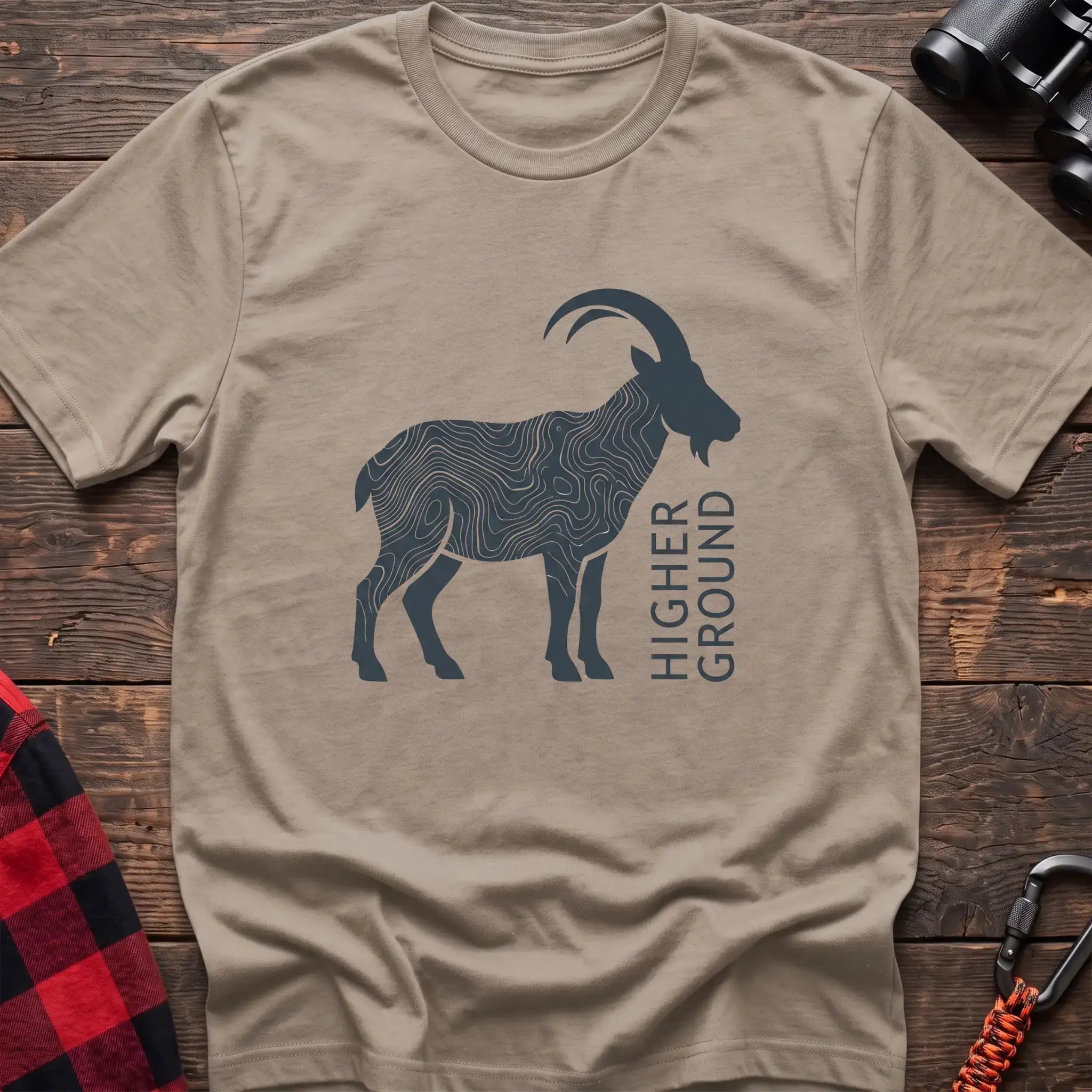Sand t-shirt with a dark silhouette of a mountain goat filled with topographic contour lines, and the vertical text “HIGHER GROUND” printed beside it, laid flat on a wooden surface.