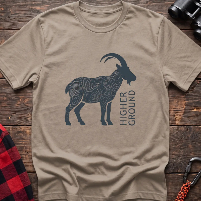 Sand t-shirt with a dark silhouette of a mountain goat filled with topographic contour lines, and the vertical text “HIGHER GROUND” printed beside it, laid flat on a wooden surface.